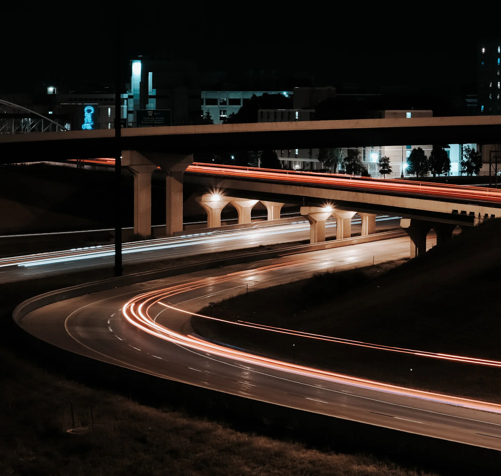 Nighttime view of highway overpasses with light trails from moving vehicles, city buildings, and illuminated signs in the background.