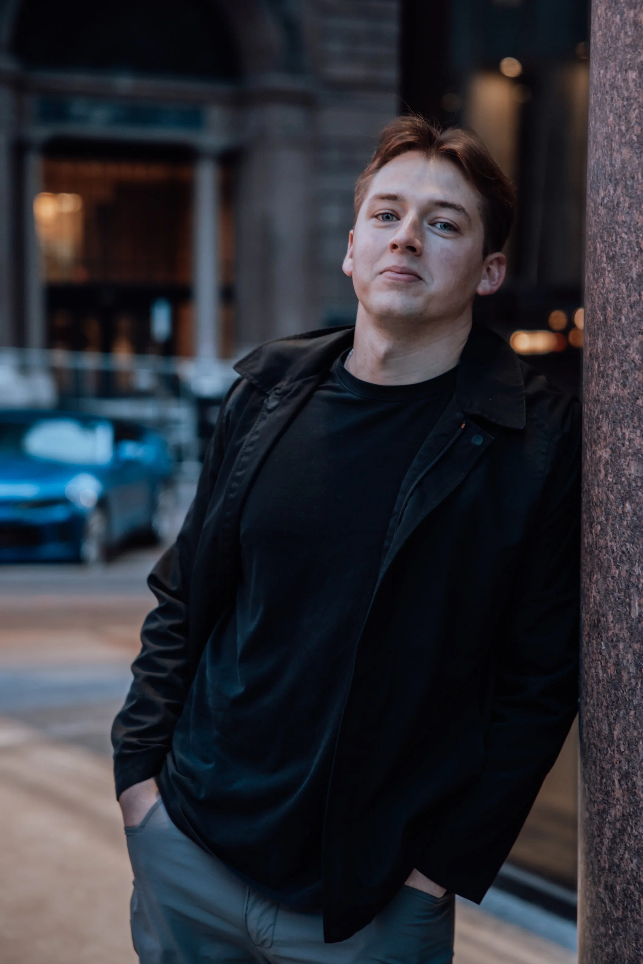 A young man with brown hair and light blue eyes is leaning against a textured brick column on a city street during dusk. He is wearing a black jacket over a black shirt and gray pants, with his left hand in his pocket. The background features parked 