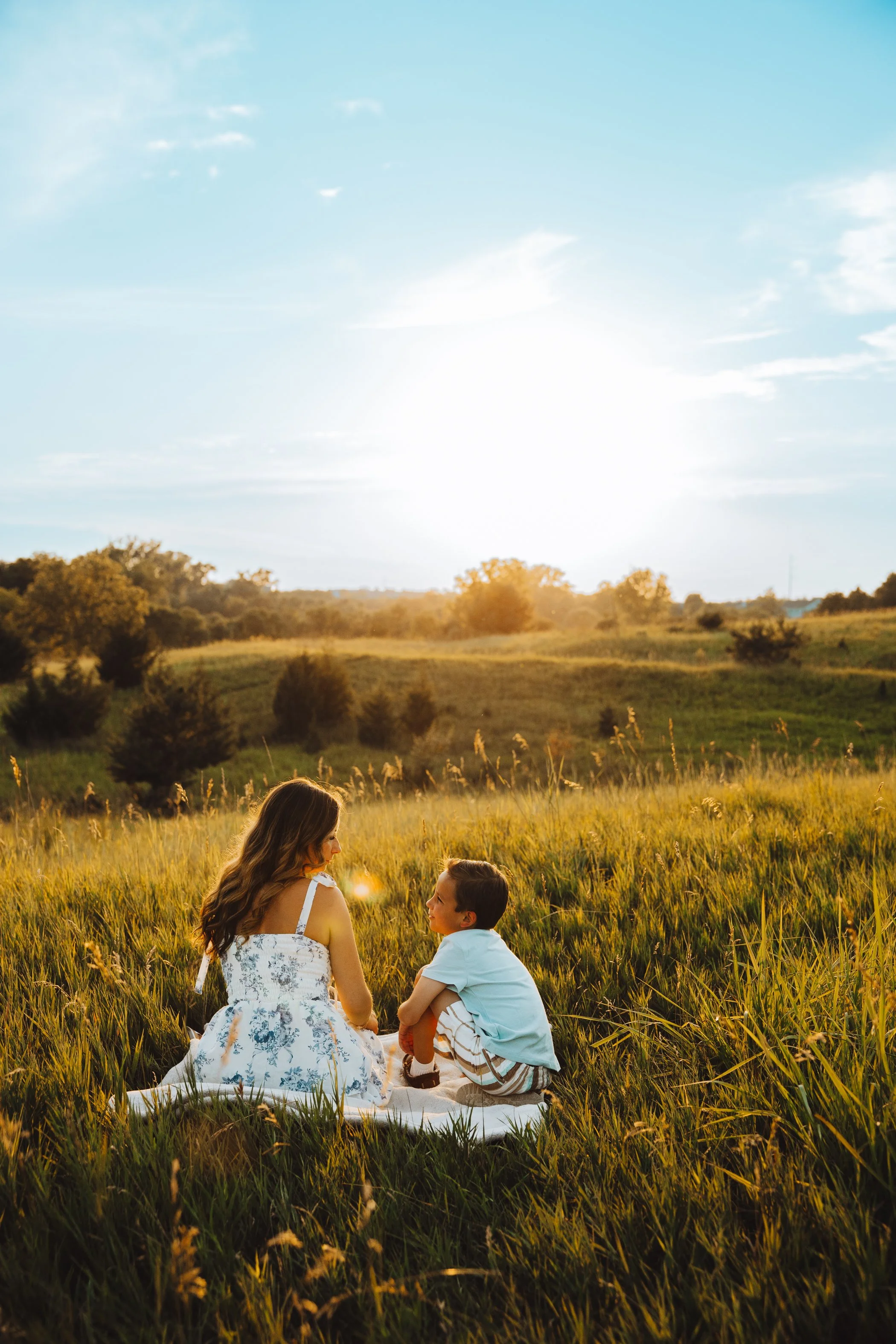 A woman and a young boy sitting on a blanket in a grassy field during sunset, with trees and a clear sky in the background.