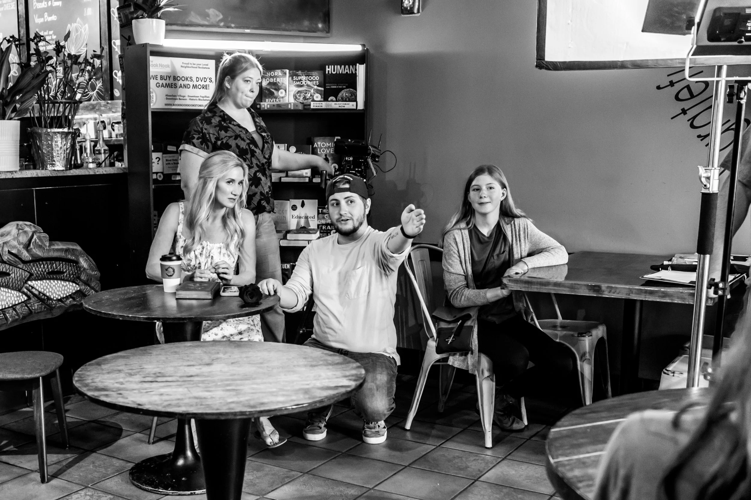 A black-and-white photo shows four people sitting at round tables in a cafe or restaurant. Behind them, a woman stands at a shelf filled with books, DVDs, and food items, operating a camera mounted on a stand. The group appears to be involved in a filming or photo shoot, with various equipment around.