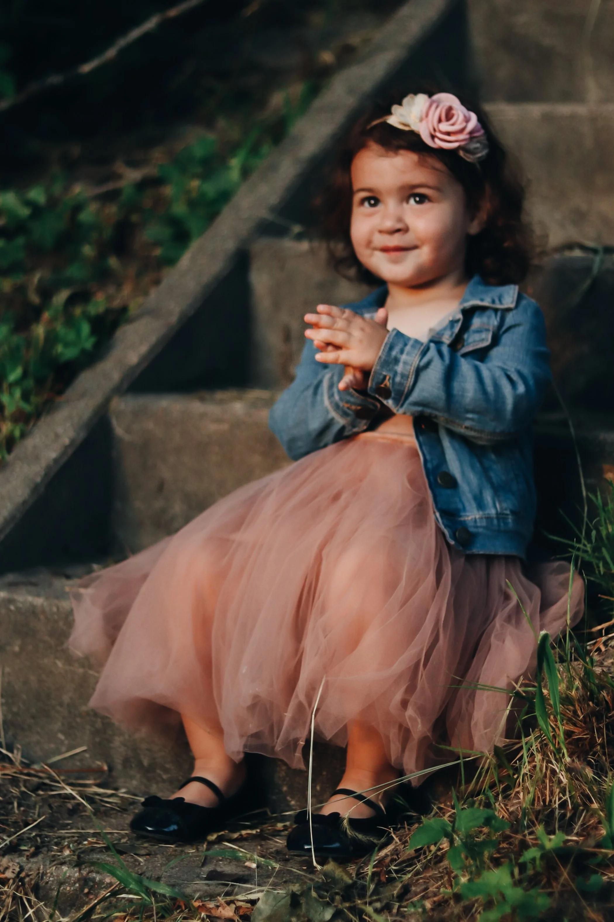 A young girl with curly hair is sitting outdoors near steps, wearing a denim jacket, a pink tulle skirt, black shoes, and a pink flower headband. She is smiling and has her hands clasped together.