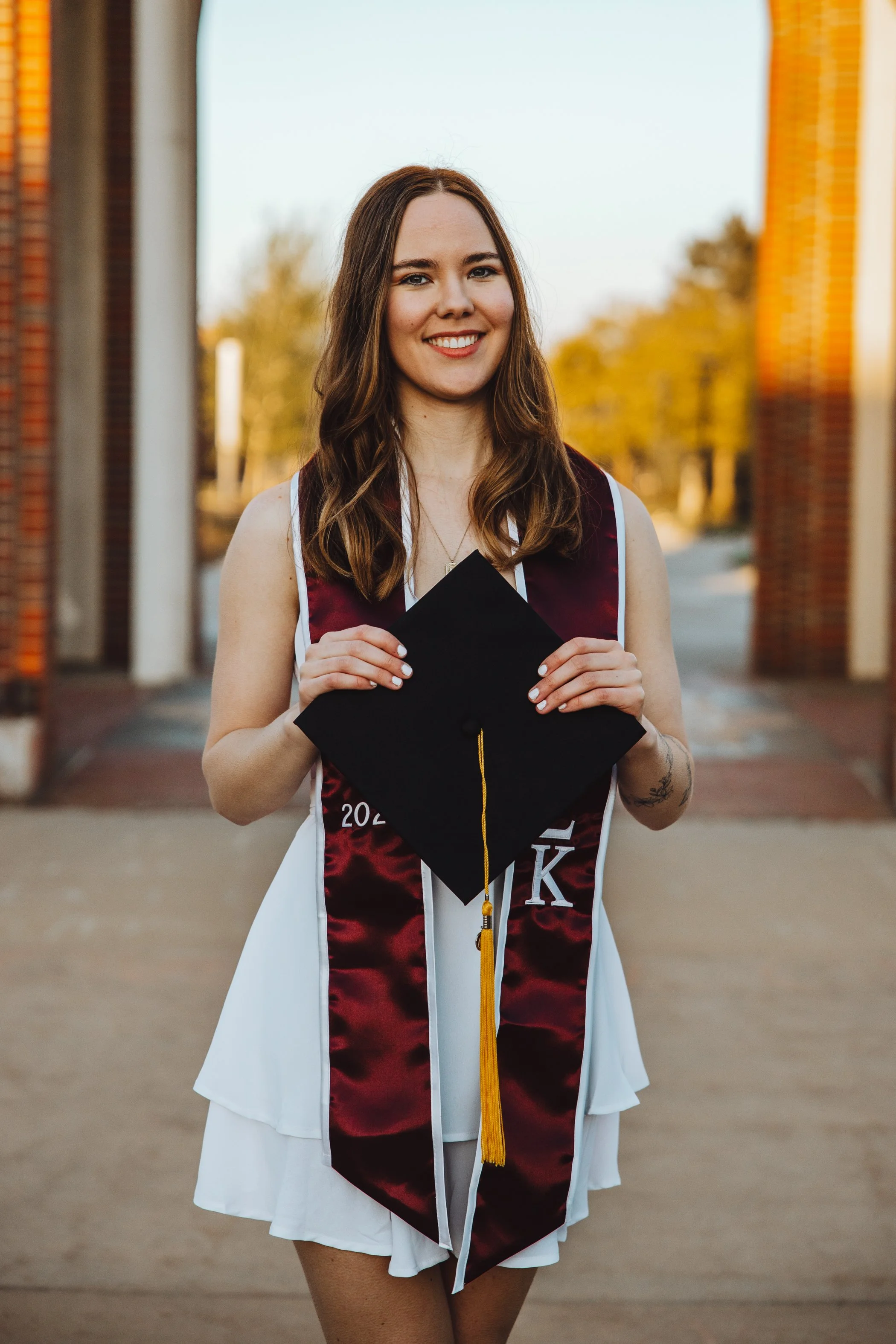 A young woman in a graduation cap and gown standing outdoors, smiling, with trees and a brick structure in the background.