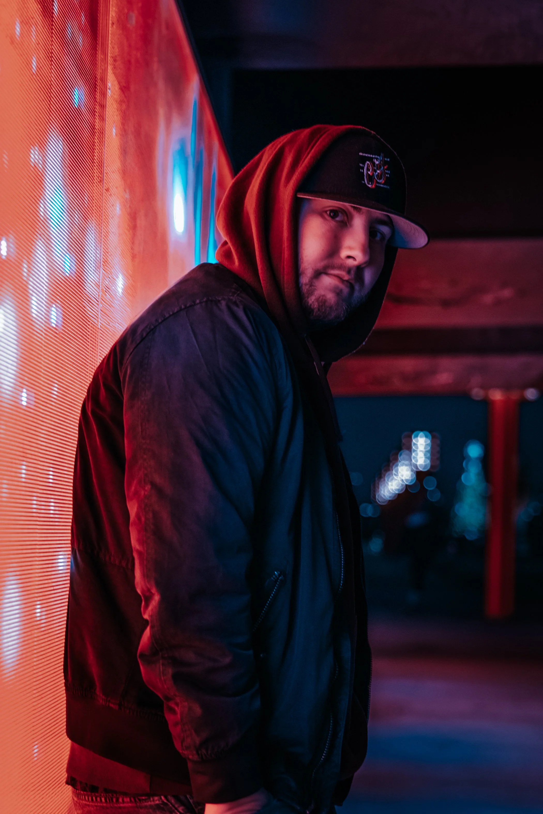 A young man with a beard wearing a black hoodie, red beanie, and black jacket stands beside a brightly lit neon sign at night. The background is dark with city lights blurred in the distance.