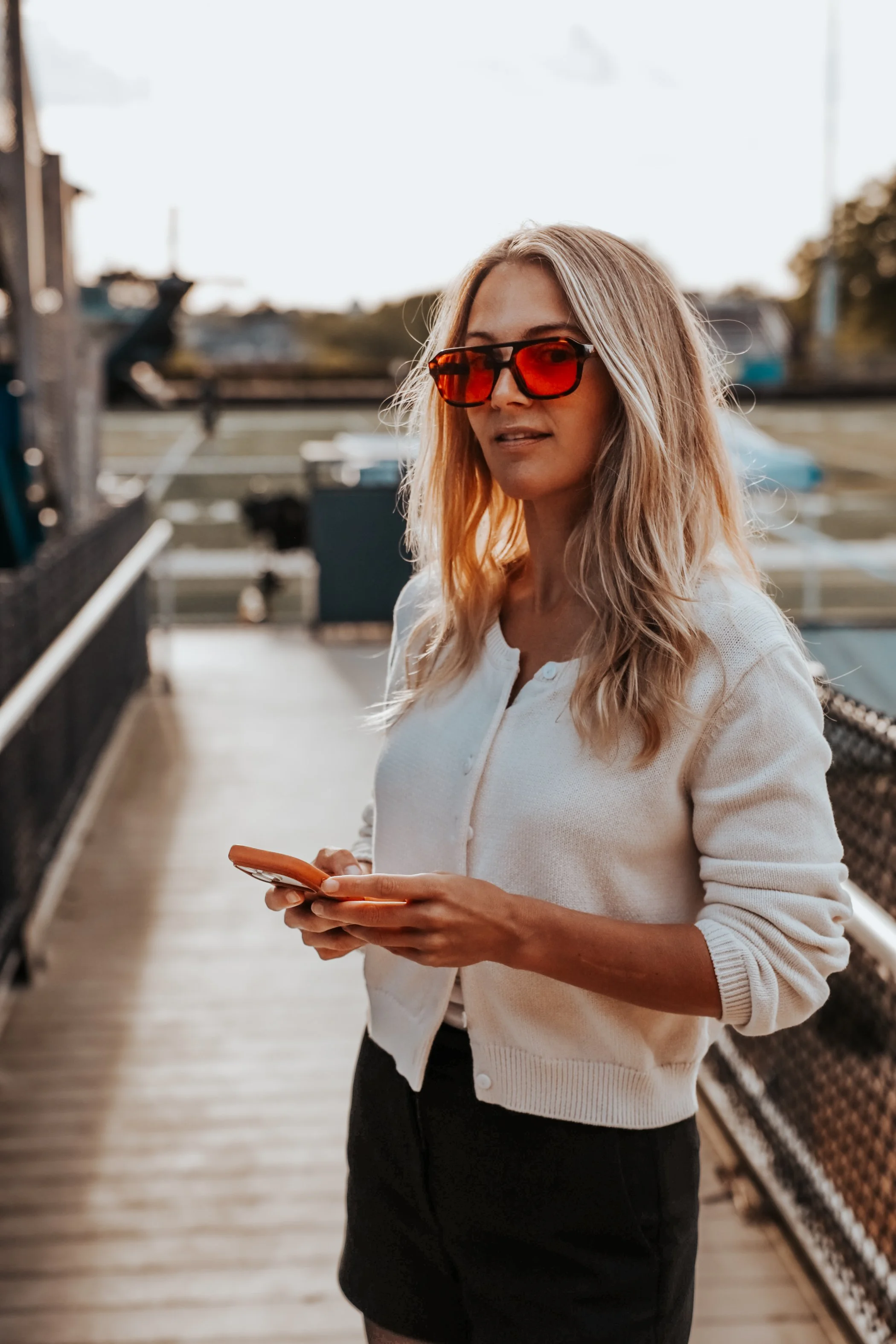 A woman with blonde hair wearing orange sunglasses and a white cardigan, holding a smartphone in her hands, standing on a wooden deck outdoors.