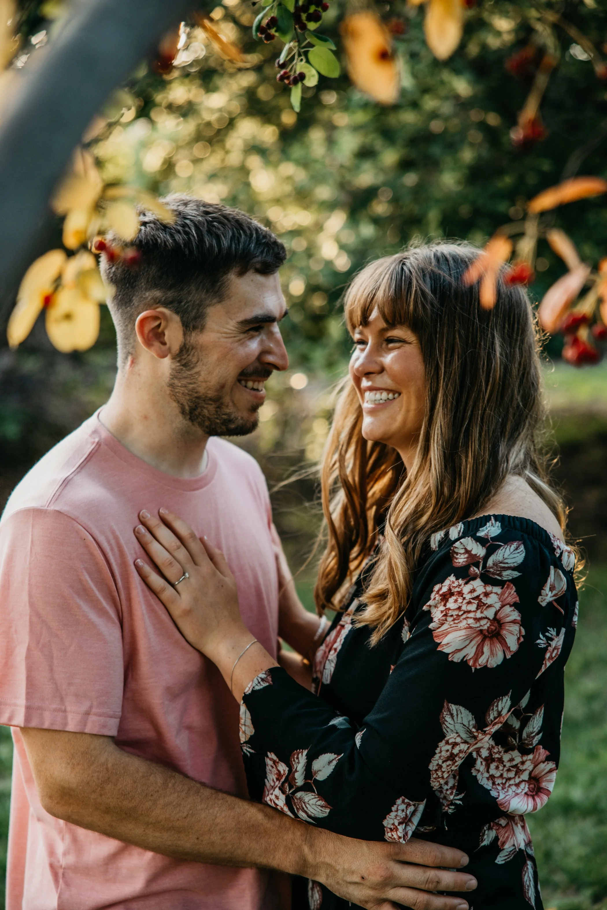 A man and woman sharing a joyful moment outdoors, smiling at each other, surrounded by trees and foliage.