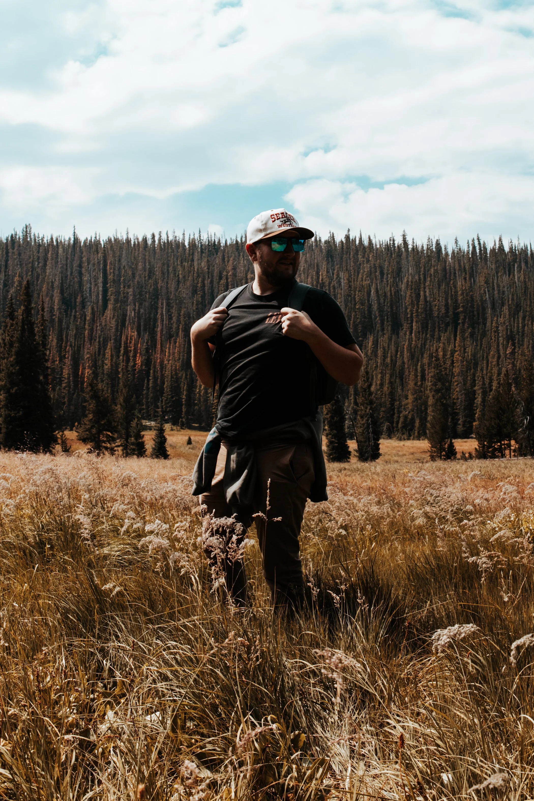 A man with a backpack and sunglasses standing in a field of tall grass with trees in the background under a blue sky with clouds.