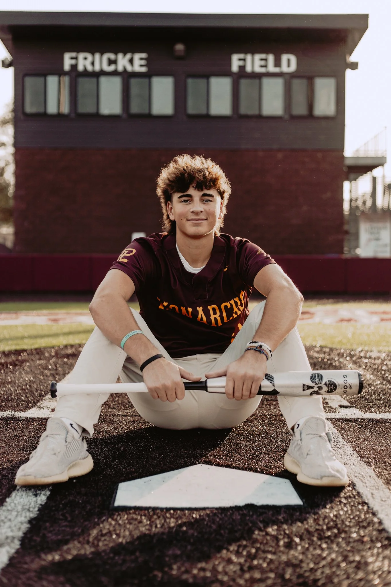 A young man with curly hair sitting on a baseball field at home plate, holding a baseball bat, with a school building that says "FRICKE FIELD" in the background.