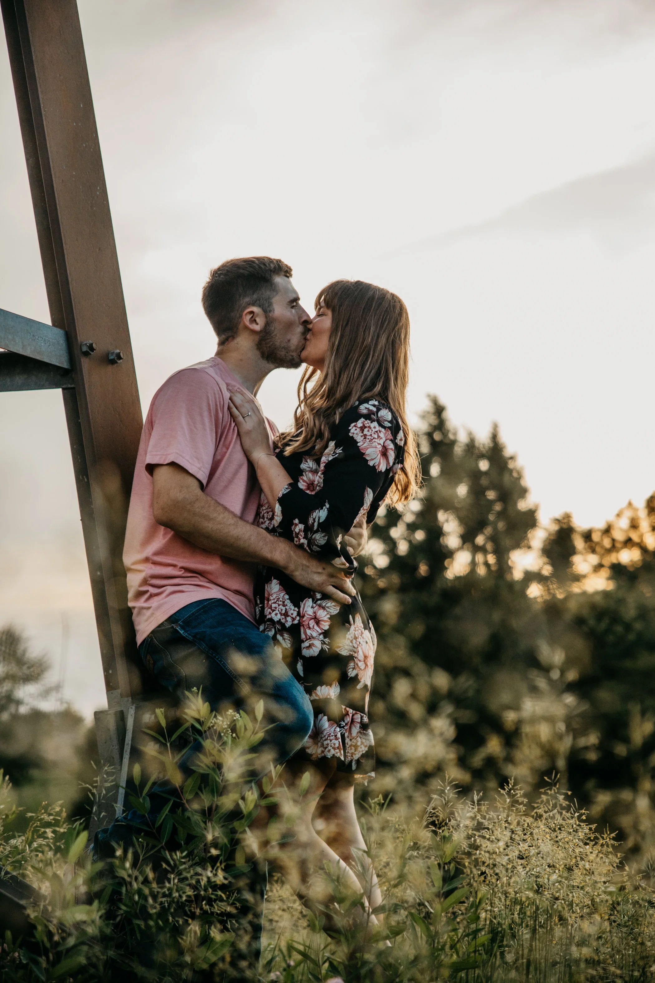A couple sharing a kiss outdoors near sunset, with trees in the background and plants in the foreground.