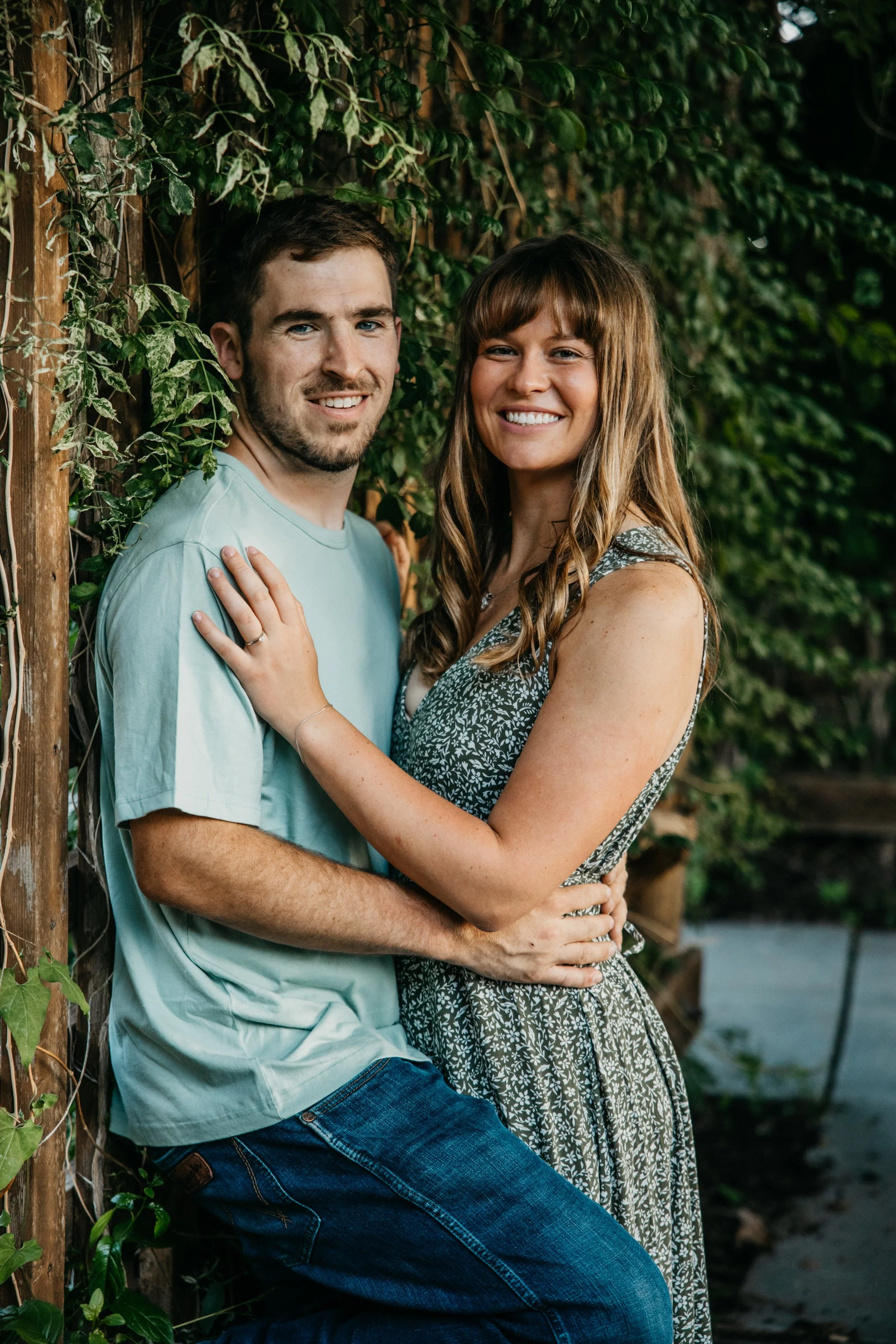 A smiling man and woman embracing outdoors near a wooden fence covered in green ivy.