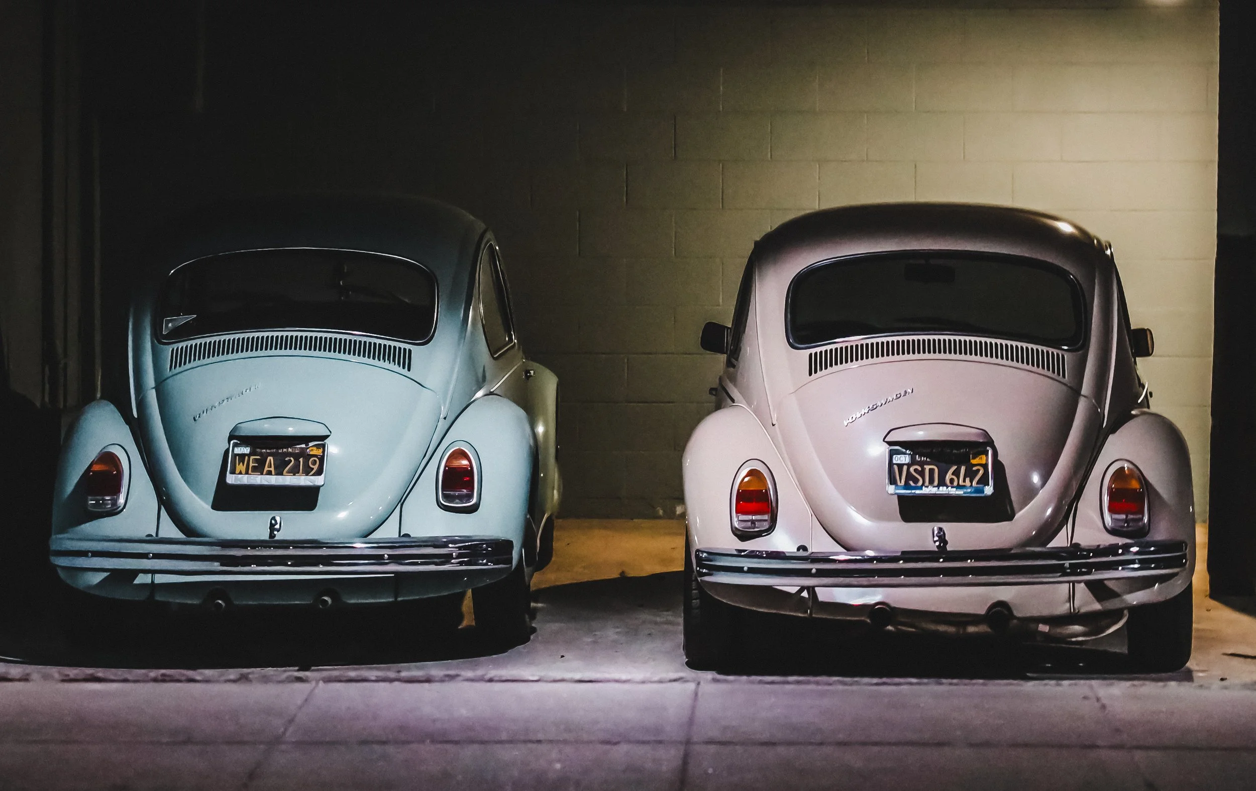 Two vintage Volkswagen Beetle cars parked side by side in a dimly lit garage. The car on the left is light blue, and the one on the right is pastel pink. Both cars are facing away from the camera, showing their rear ends with license plates and simil