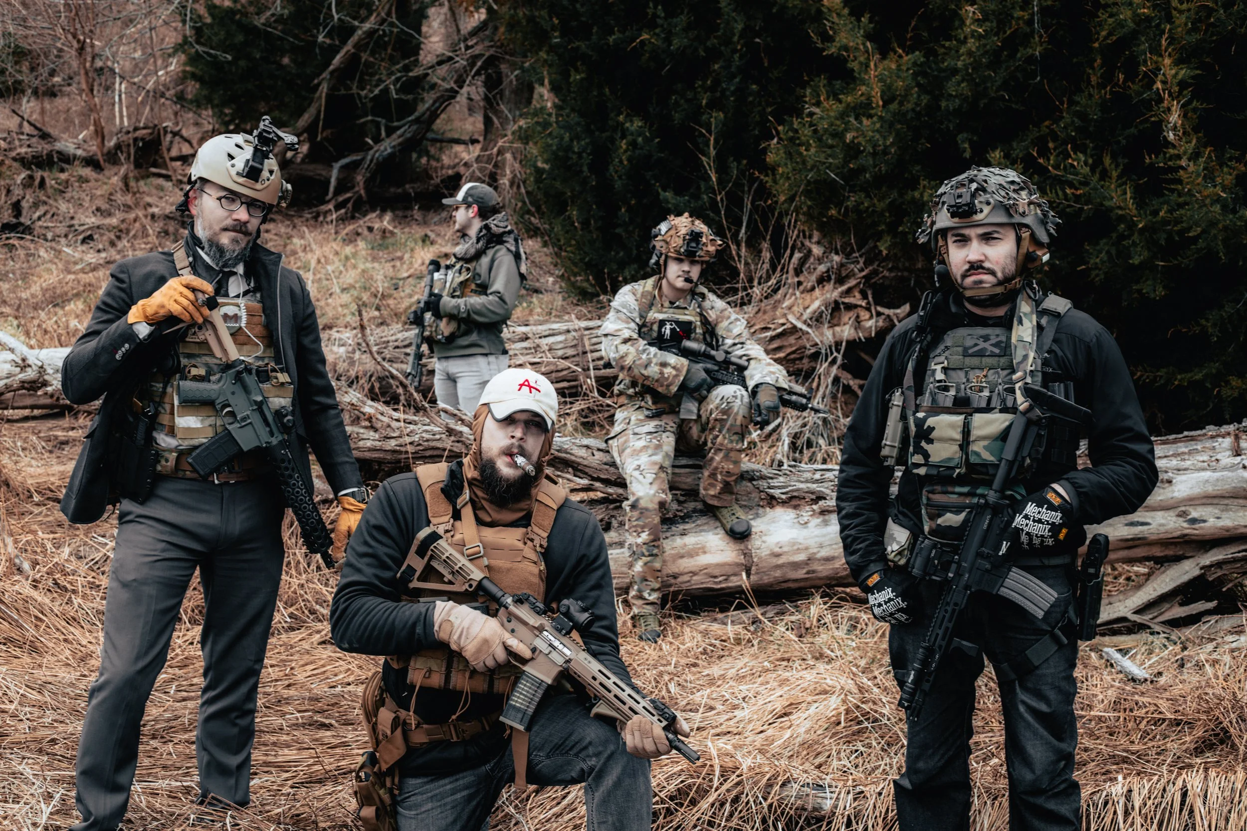 A group of five men dressed in tactical gear, posing outdoors in a wooded area with fallen tree branches. Four of them are holding firearms, and all are wearing helmets and outdoor clothing. One man is smoking a cigarette.