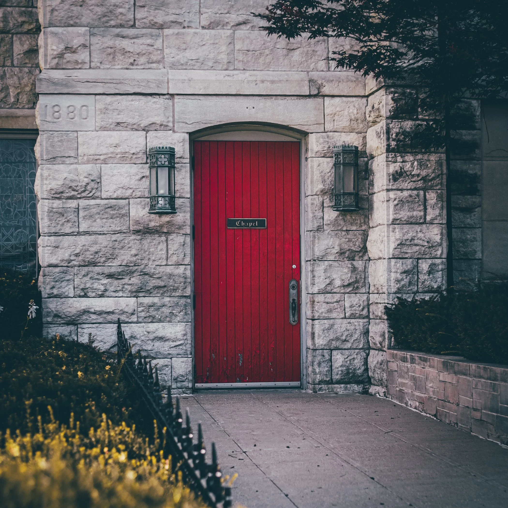 Stone chapel entrance with red door, stone walls, lantern-style lights, and a small sign that reads "Chapel," surrounded by yellow flowers and greenery.