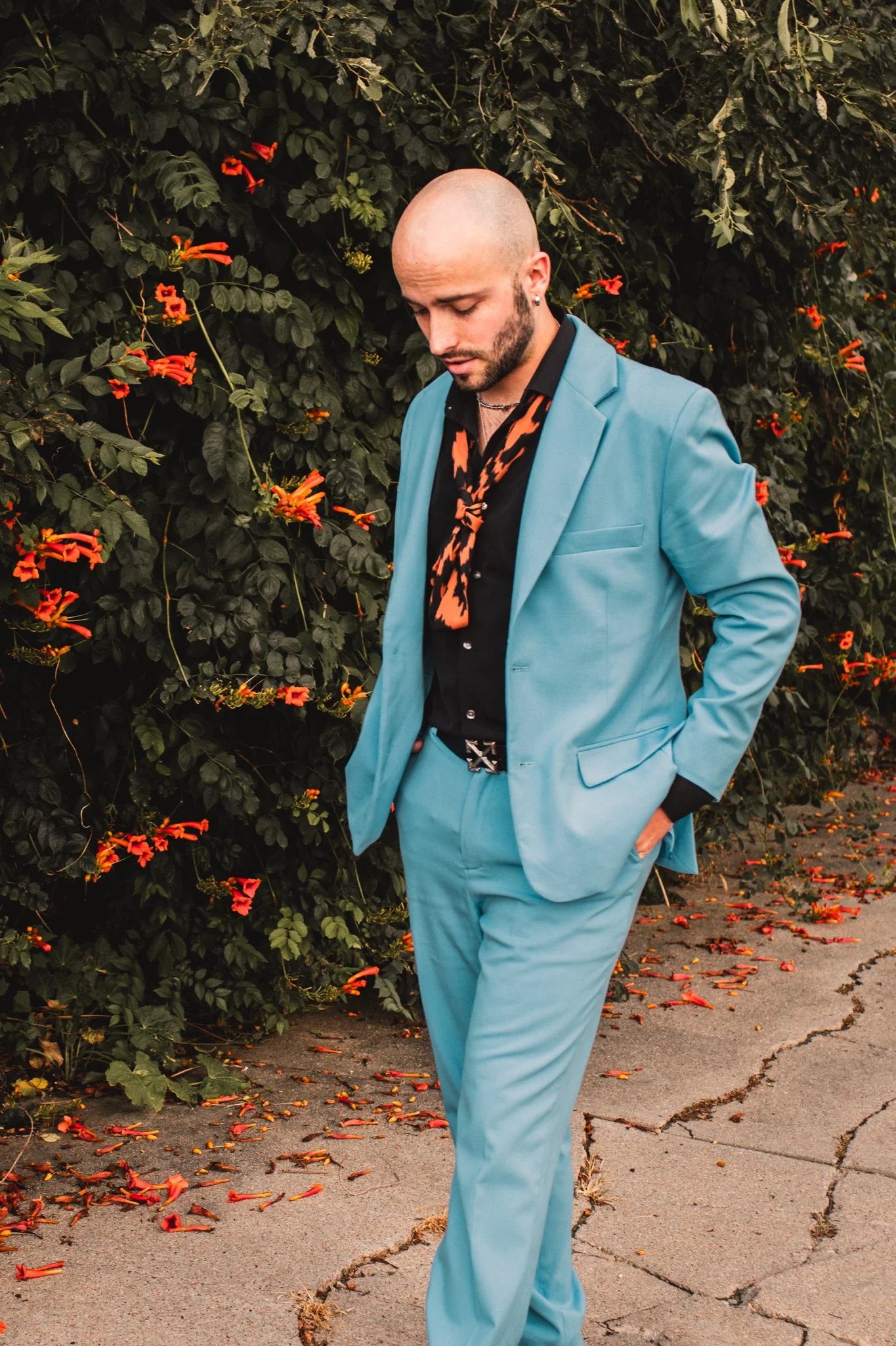 A man in a light blue suit and black shirt with orange patterns, with a bald head and beard, stands with his hands in his pockets in front of a bush with orange flowers. The ground has fallen orange flower petals and cracks in the concrete.