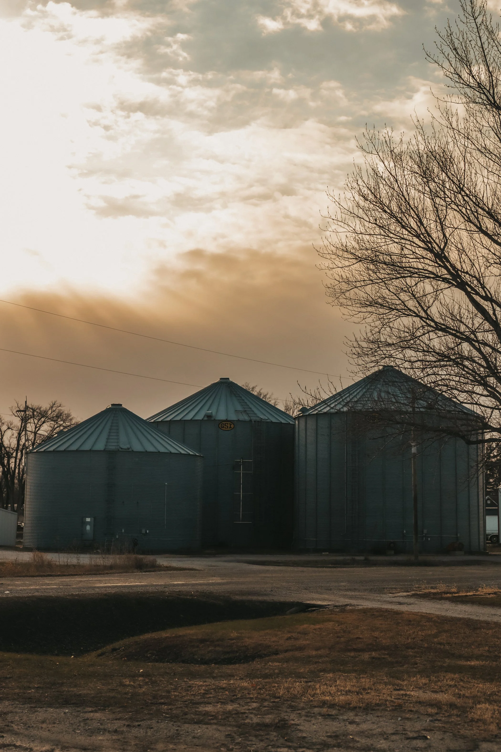 Three large metal grain silos with conical roofs, set against a cloudy sky during sunset, with leafless trees nearby and an empty dirt area in the foreground.