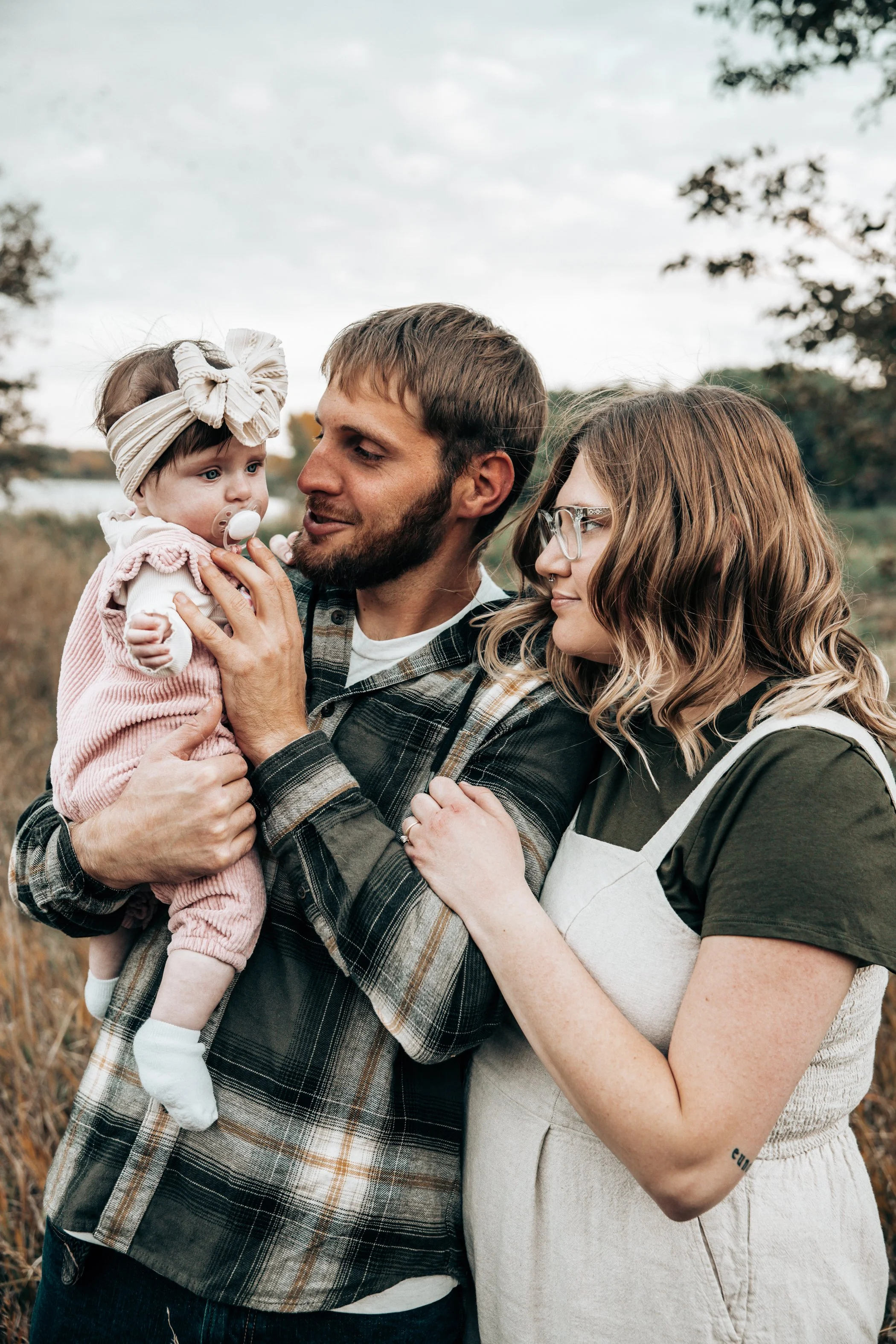 A family of three outdoors in a field with trees in the background. The father is holding a baby girl with a pacifier, and the mother is standing beside them, touching the father's arm. The baby is wearing a pink outfit and a headband, while the pare