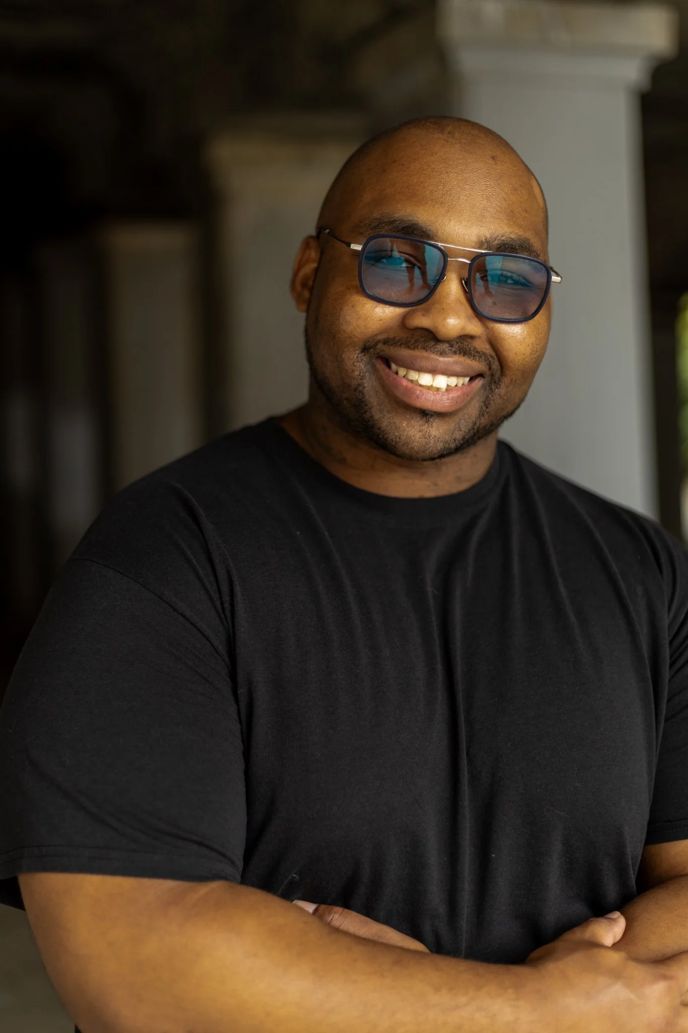 A smiling man wearing sunglasses and a black t-shirt, standing outdoors with his arms crossed.
