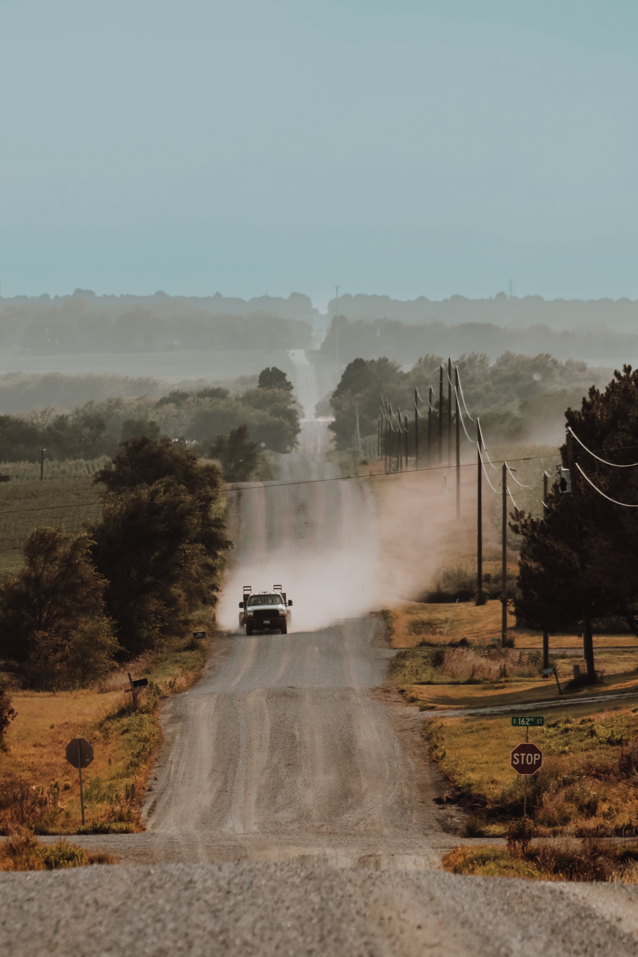 A rural dirt road with a truck driving on it, surrounded by trees and fields, with power lines on the right and a stop sign at the intersection.
