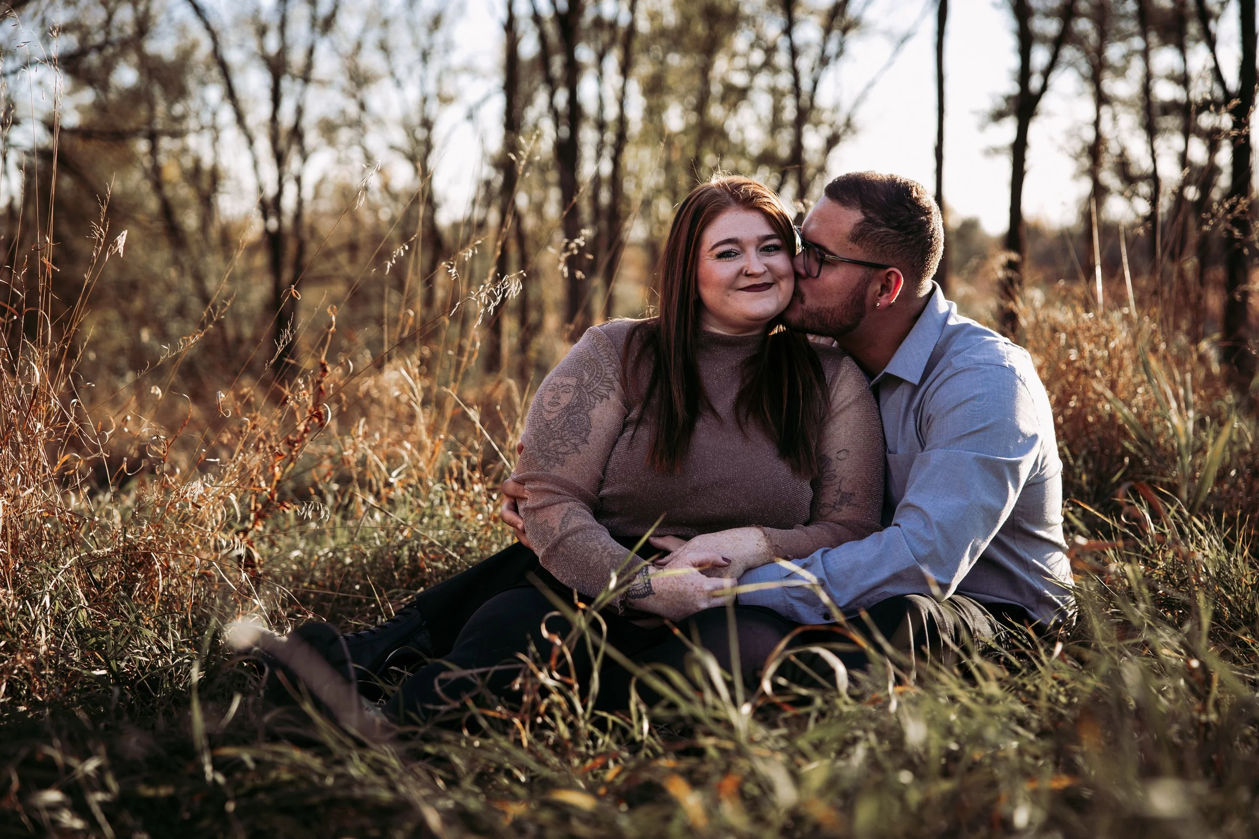 A couple sits on the ground in a field with tall, dry grass, embracing and smiling; the man is kissing the woman on the cheek while she smiles and looks at the camera. They are surrounded by trees with sunlight filtering through.