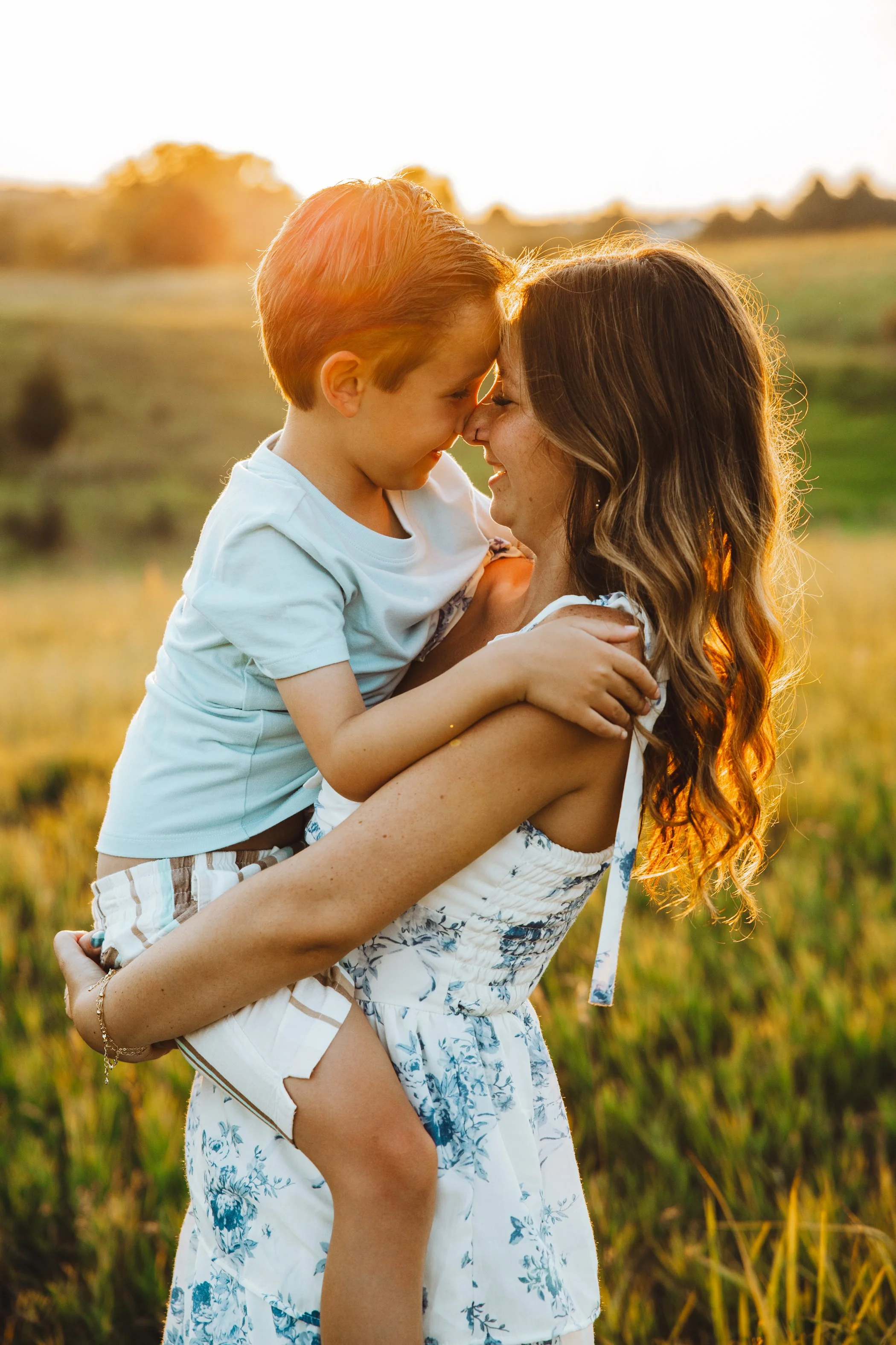 A woman with long wavy hair lifting a young boy with red hair in her arms outdoors at sunset, both touching foreheads and smiling in a field of tall grass.