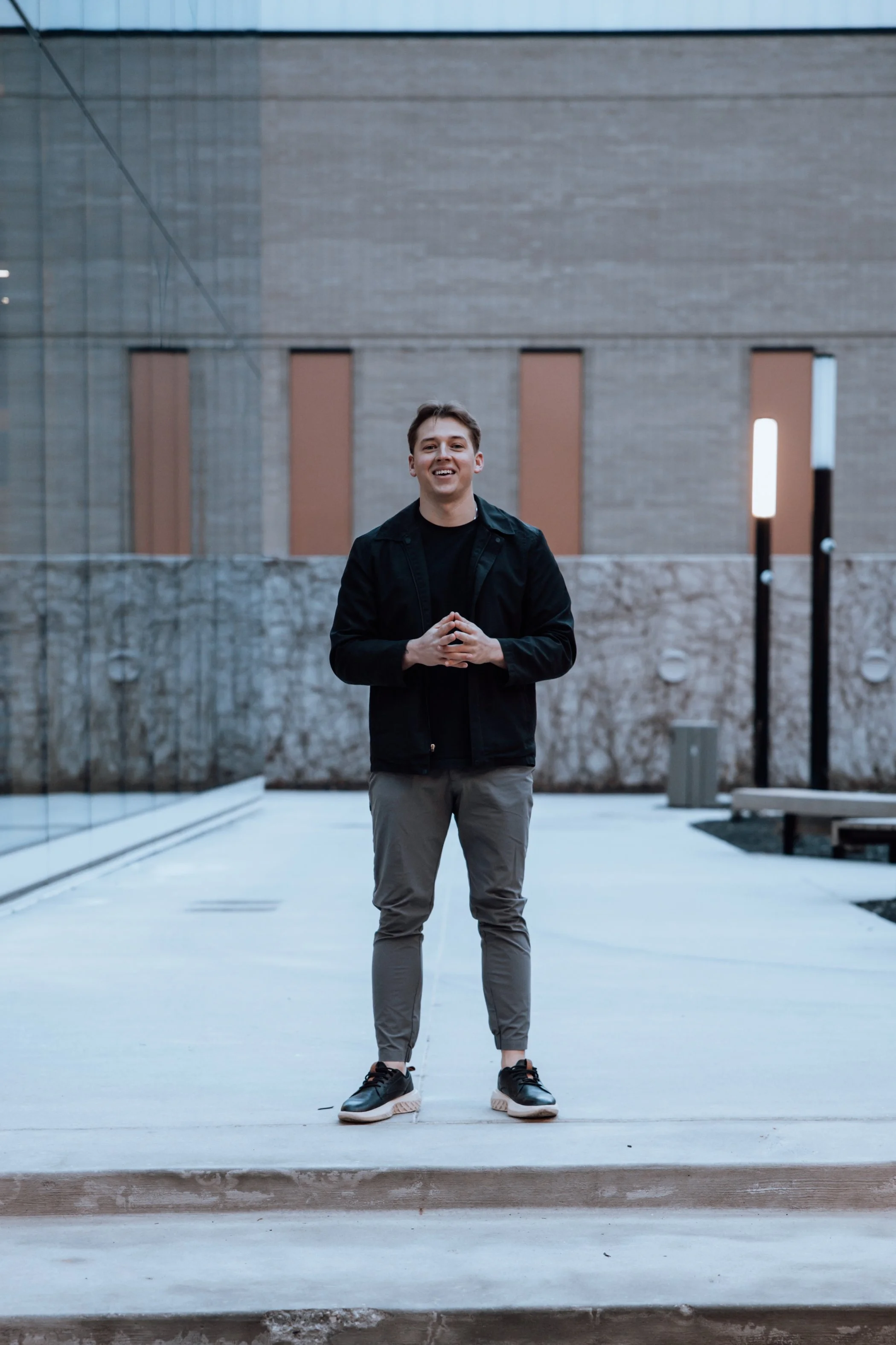 A young man stands outdoors on a concrete pathway, smiling and looking at the camera with his hands clasped in front of him. He is wearing a black jacket, gray pants, and black sneakers. The background features a modern building with large glass wind