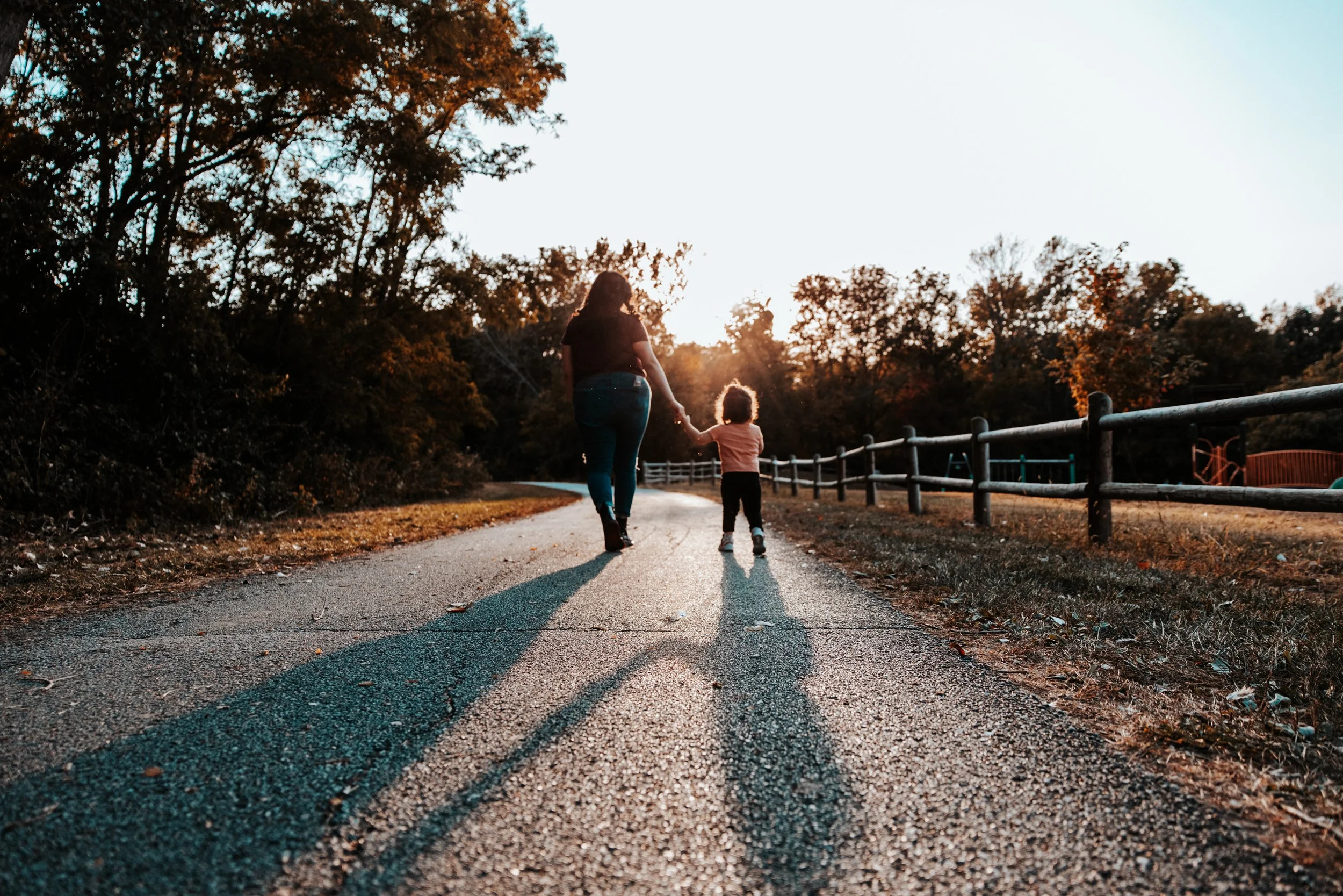 A woman and child walking hand in hand on a gravel path during sunset, surrounded by trees and a wooden fence.