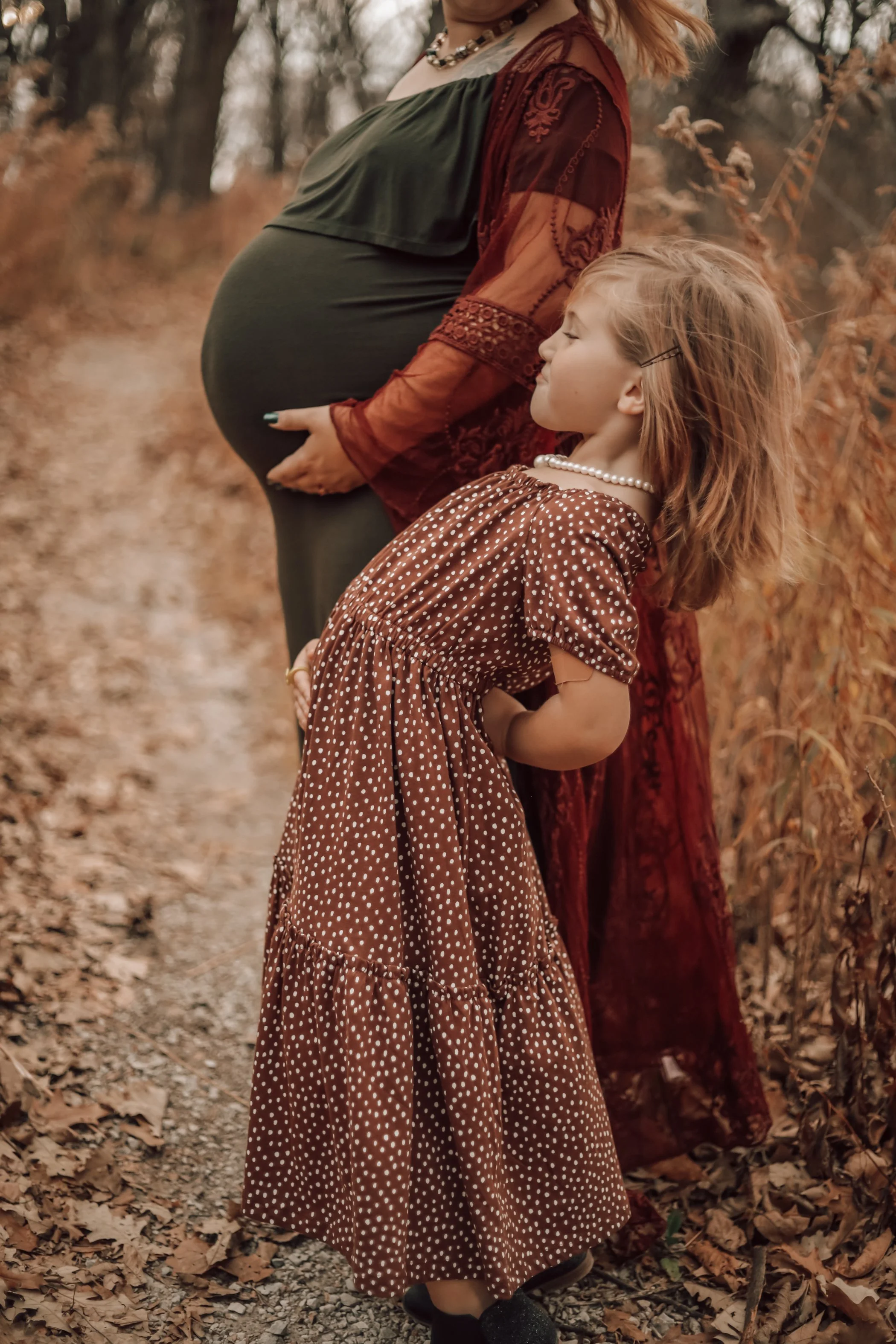 A pregnant woman and a young girl with red hair standing outdoors in a wooded area with fallen autumn leaves.