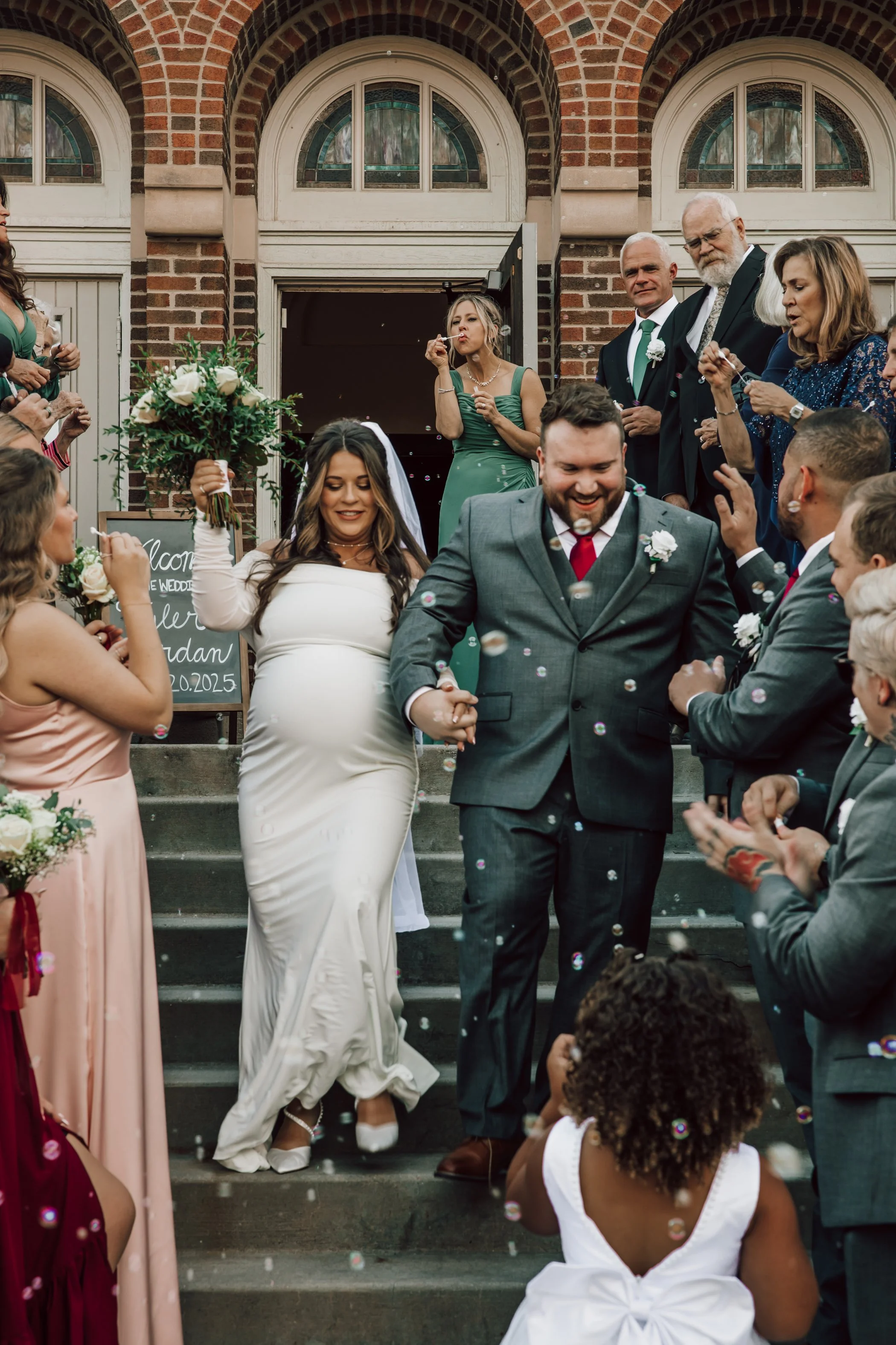 Wedding celebration on the steps of a brick building with guests throwing confetti and blowing bubbles, including a woman in a white wedding dress, a man in a gray suit, and various guests clapping and smiling.