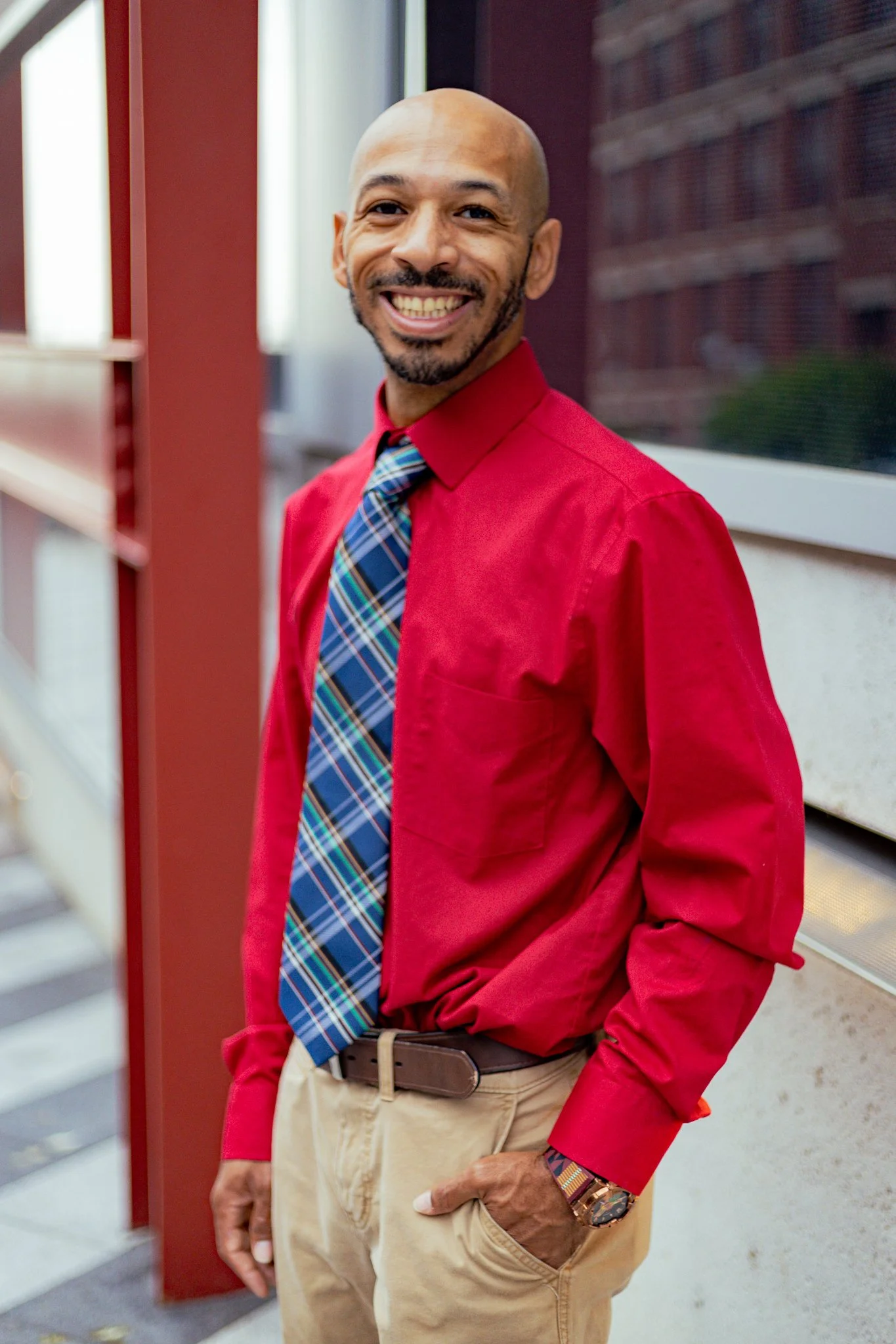 A man smiling, wearing a red dress shirt, a plaid tie, and beige pants, standing outside near a window and a brick building.