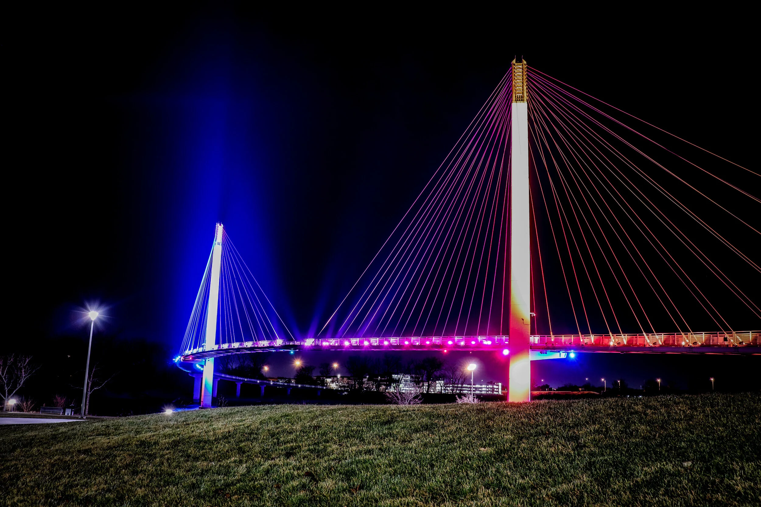 Night view of a brightly lit modern suspension bridge with colorful LED lights illuminating the cables and roadway, against a dark sky.