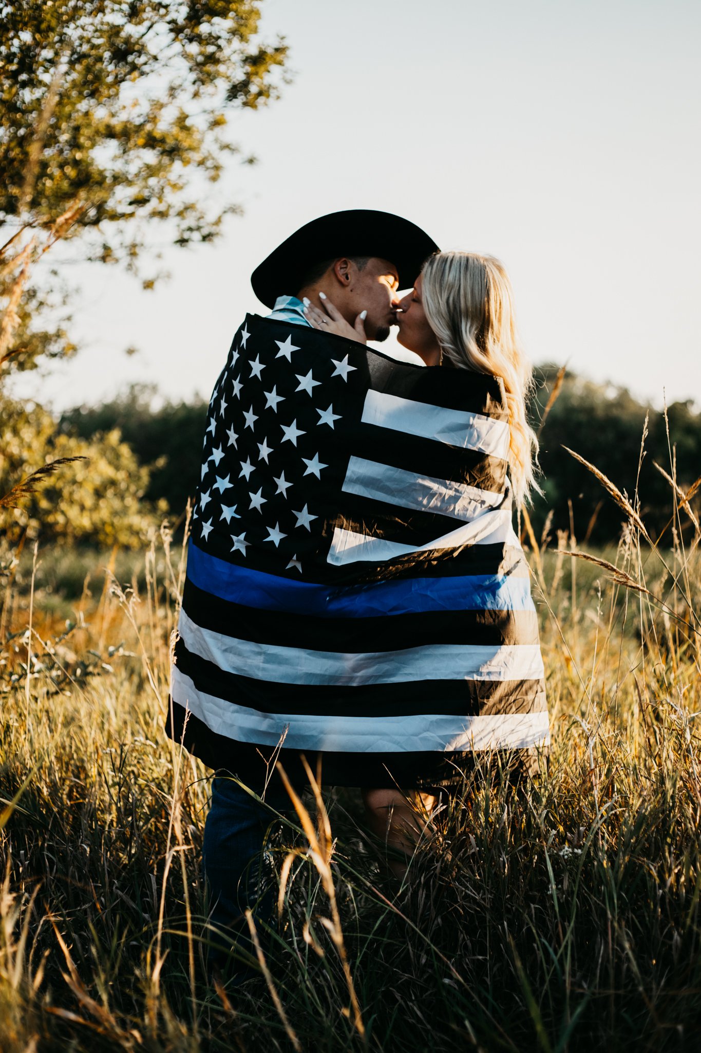 A couple is kissing outdoors in a field during sunset. The man is wearing a cowboy hat and a jacket with an American flag design. The woman has long blonde hair. They are surrounded by tall grass and trees.
