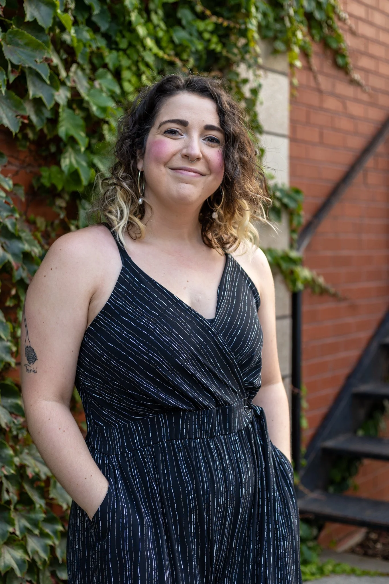 A woman with curly hair, wearing a black dress with white pinstripes, standing outdoors in front of a brick wall with green ivy, smiling at the camera.