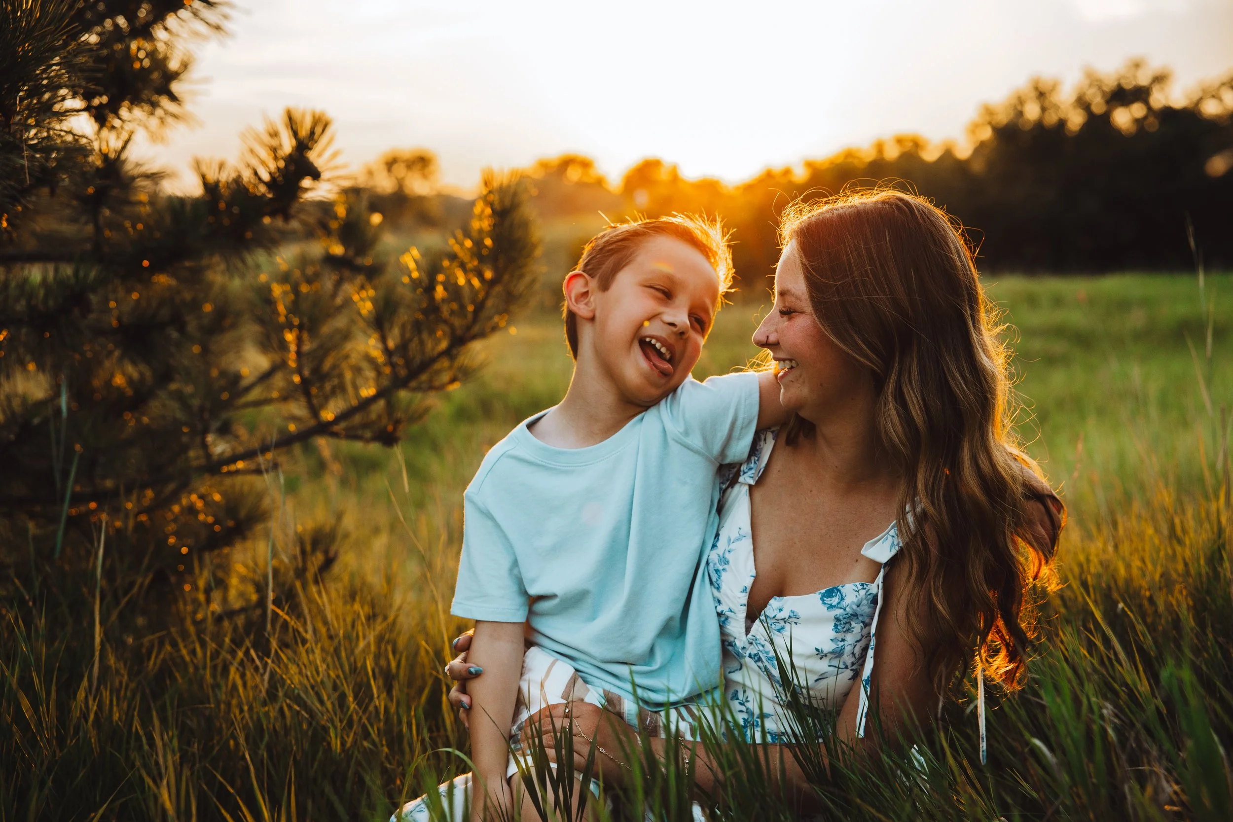 A woman and a young boy sharing a joyful moment outdoors during sunset, with trees and tall grass in the background.
