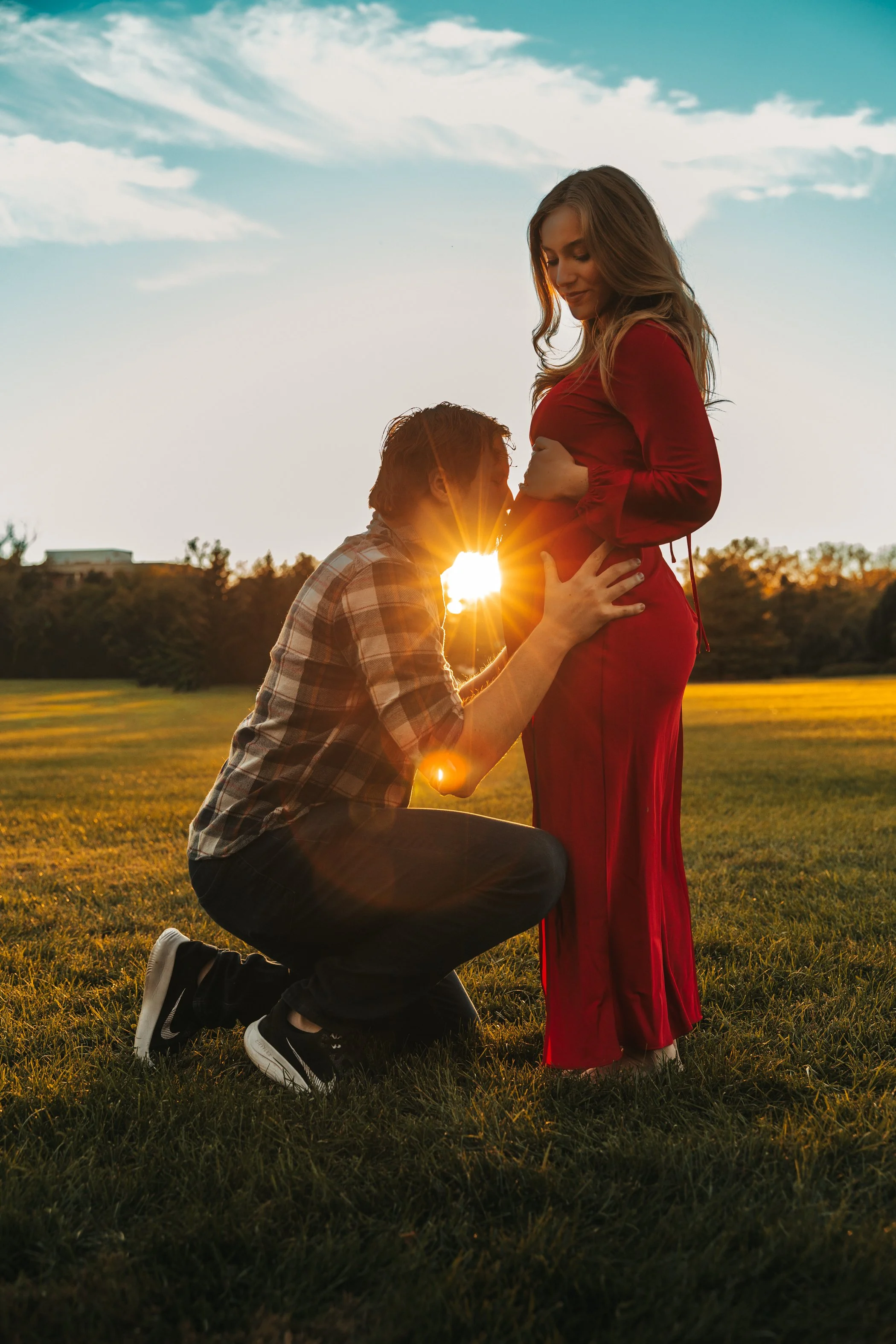 A man kneeling and a pregnant woman standing in a park during sunset, with the man kissing her belly.