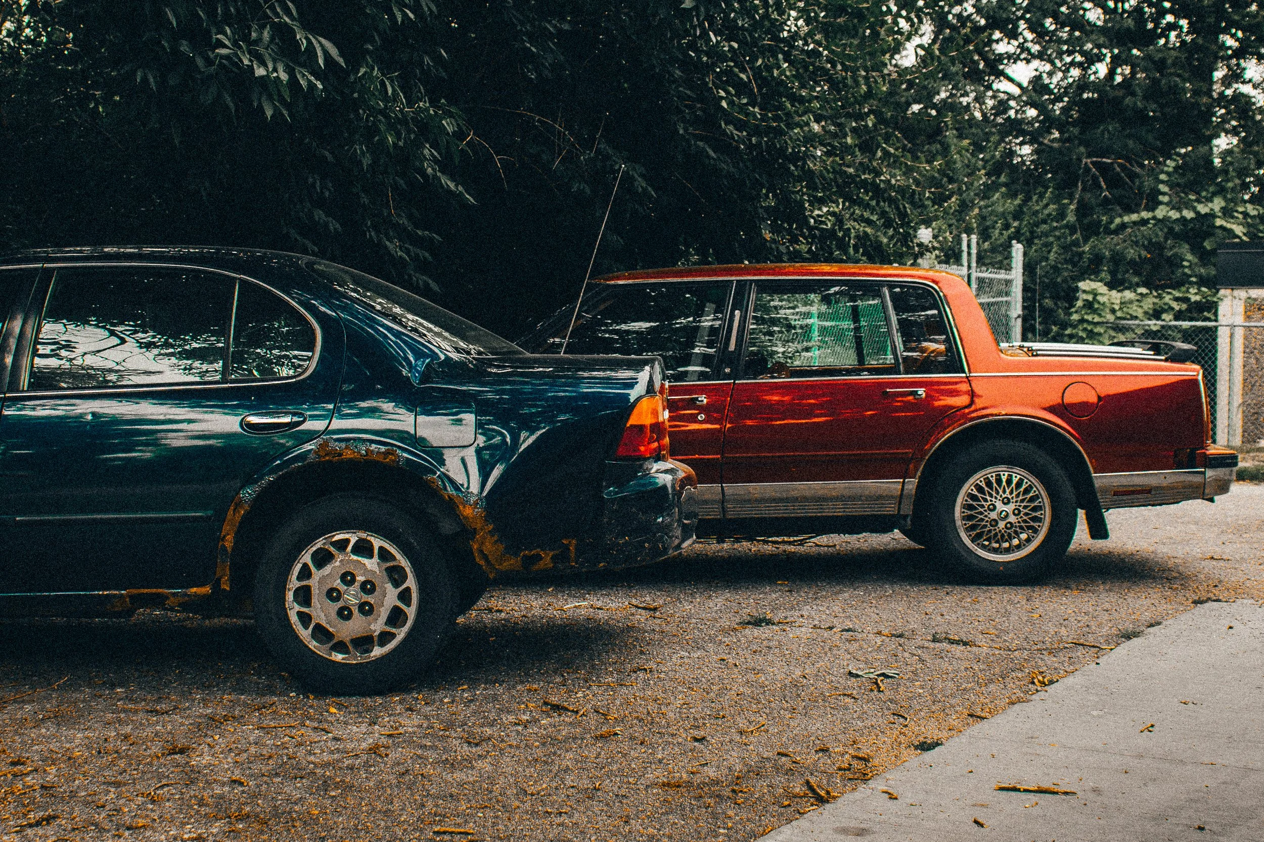 Two cars parked outdoors near a chain-link fence with trees in the background; one black sedan and one red and black pickup truck, both showing signs of rust.
