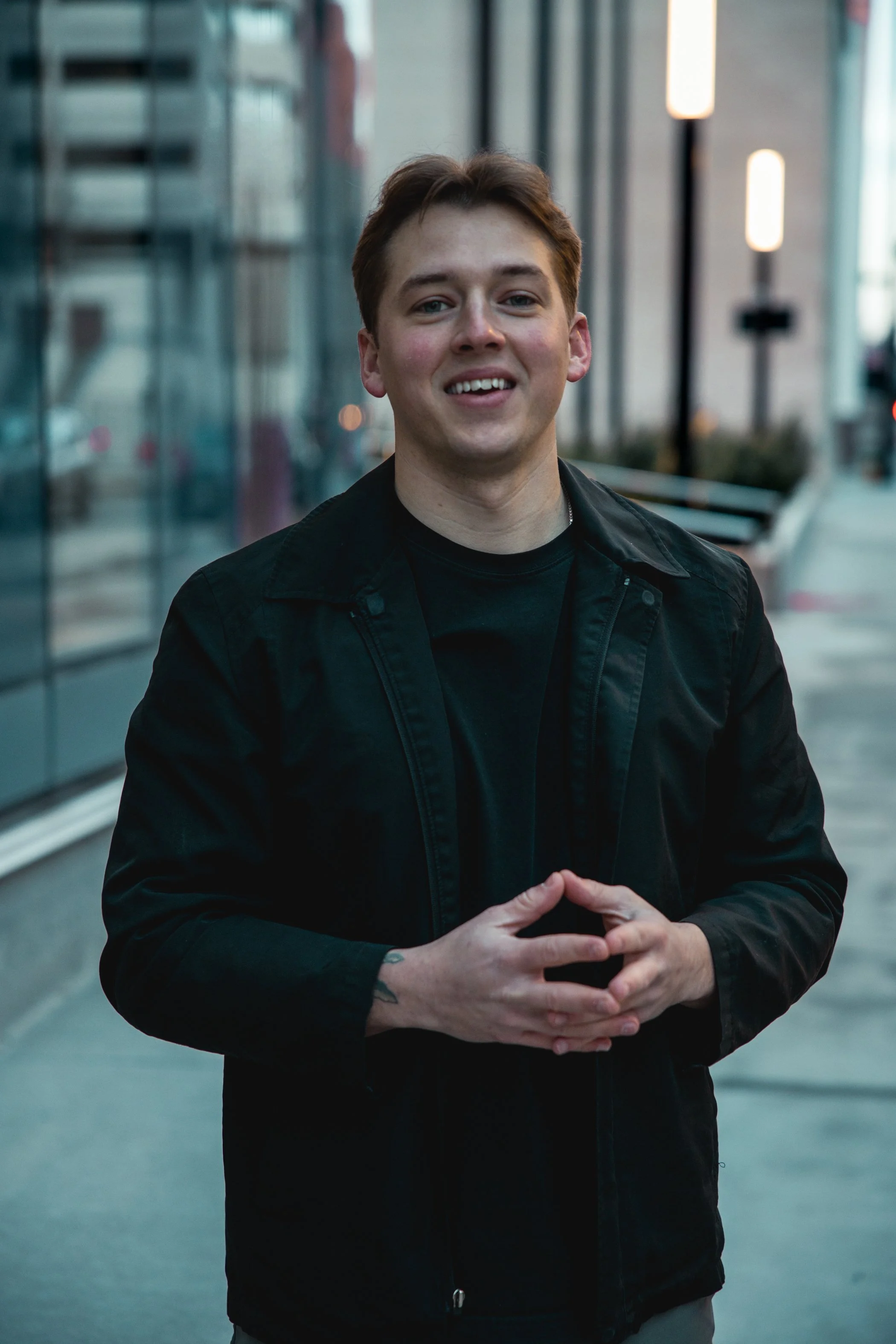 A young man wearing a black jacket with a dark shirt underneath, standing outdoors on a city sidewalk during daytime, smiling and clasping his hands together.