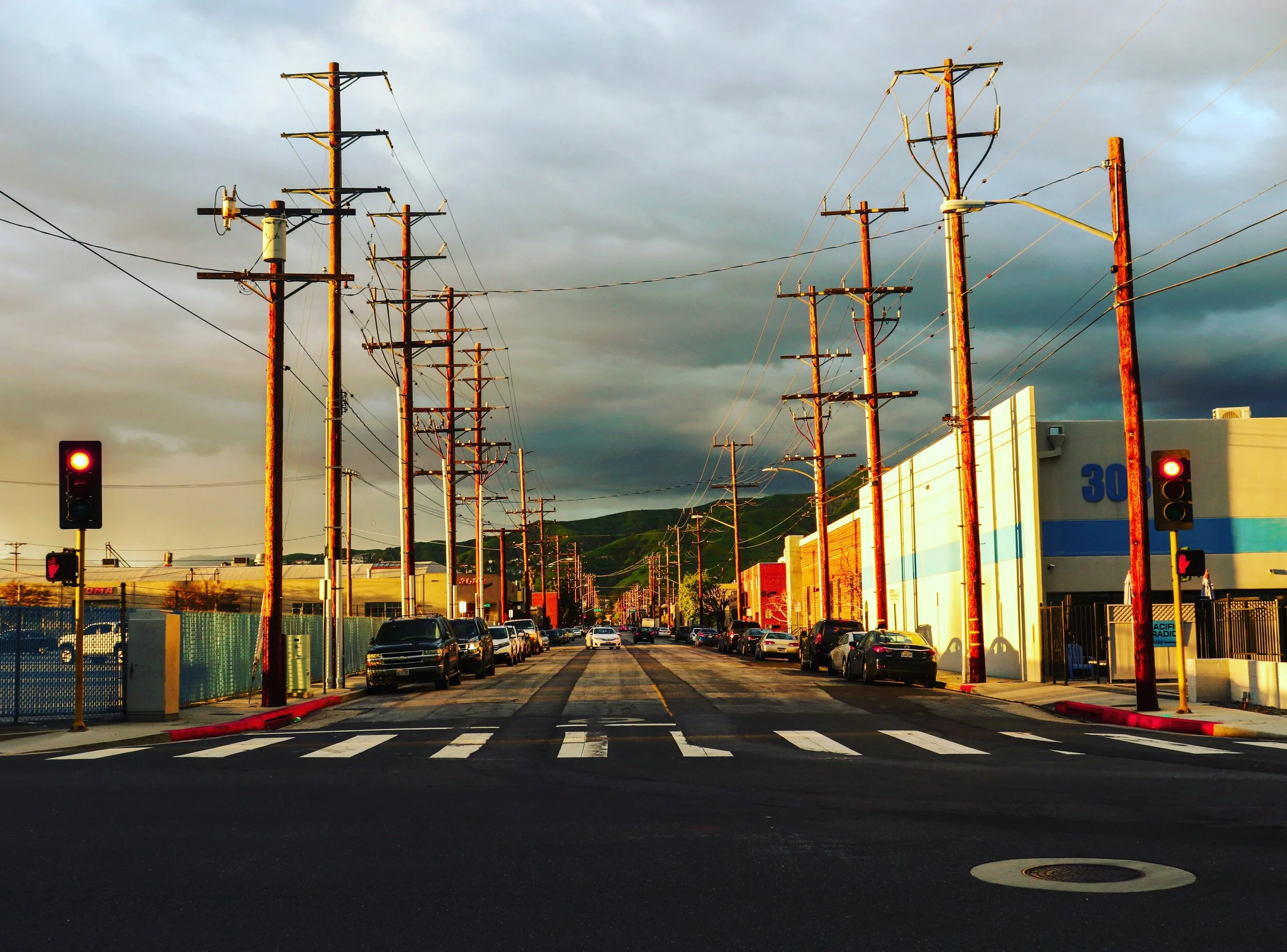 City street with power lines and poles, traffic lights, parked cars, industrial buildings, and a mountain in the background, under cloudy sky.