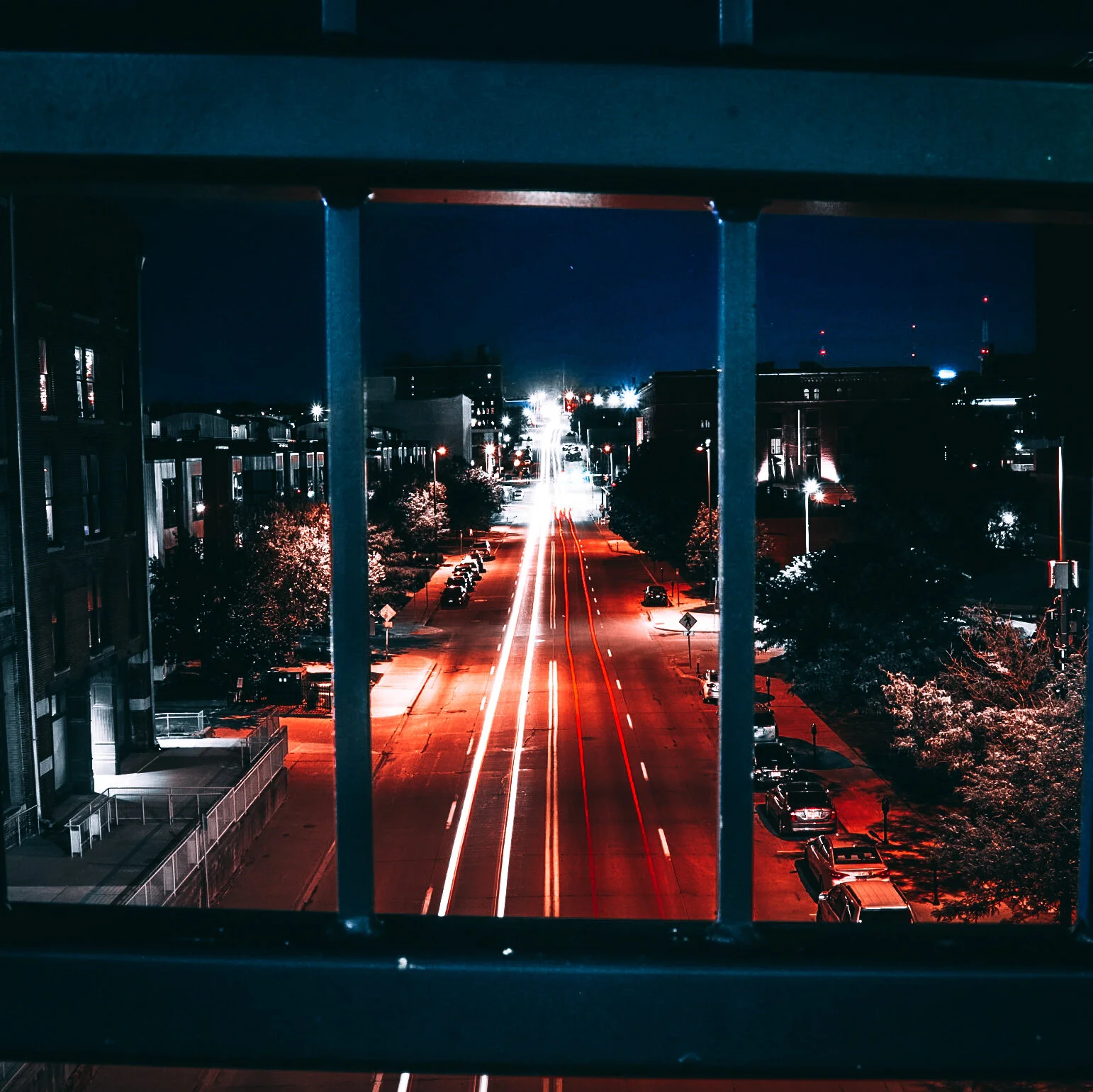 Nighttime view of a city street through window bars, with car light trails and illuminated buildings in the distance.