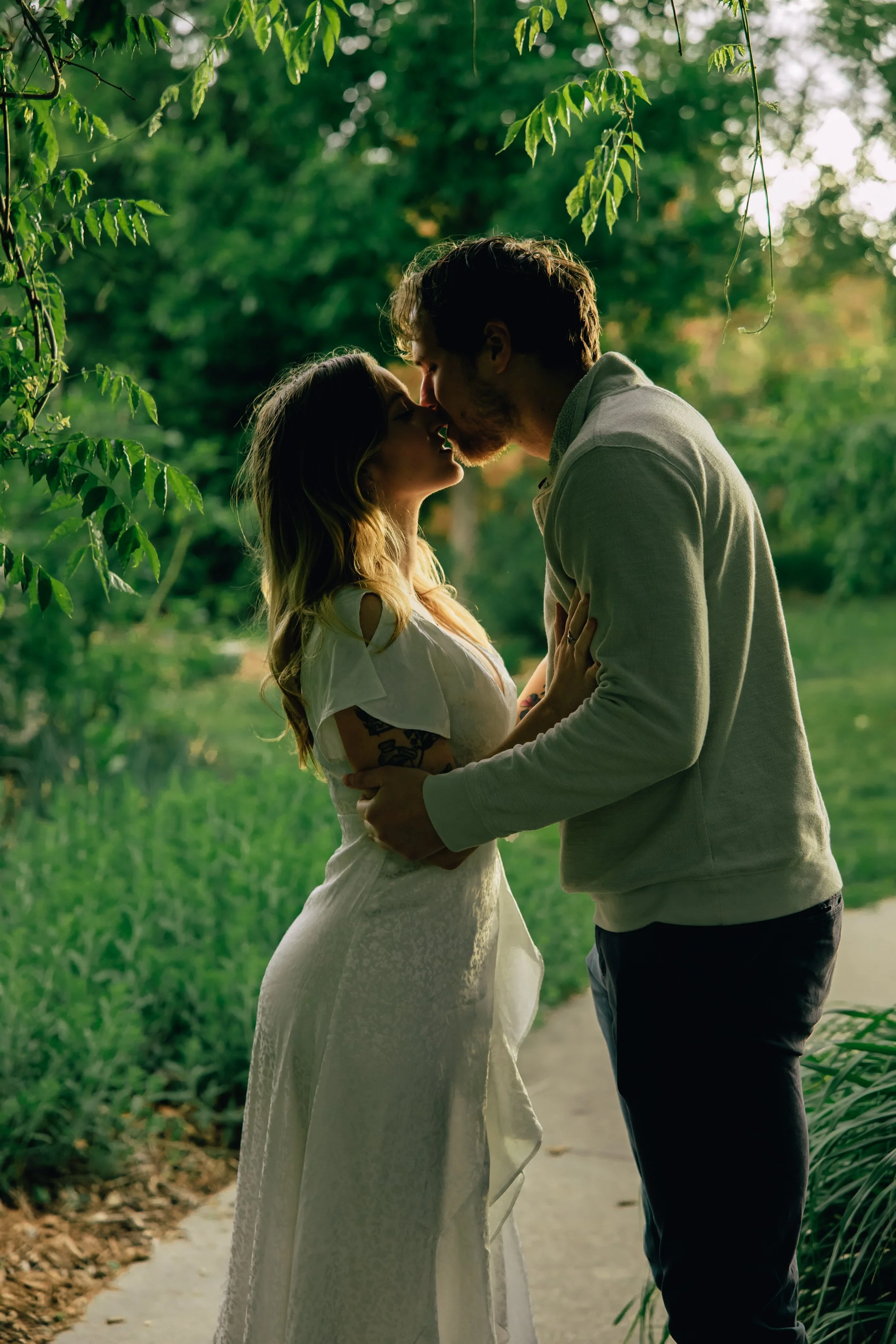 A couple stands close together under a canopy of green leaves, leaning in for a gentle kiss in a lush outdoor setting.