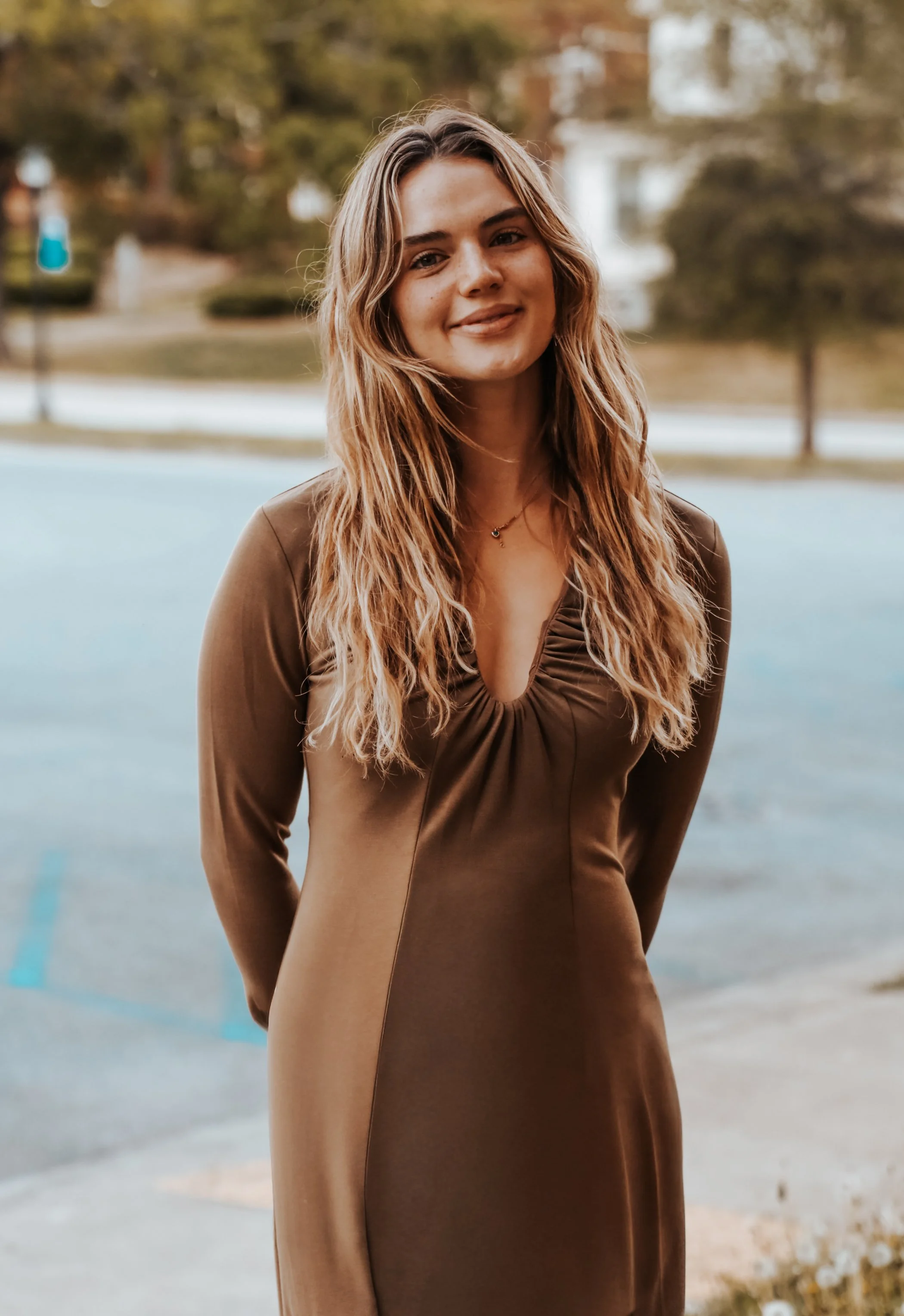 A young woman with long, wavy blonde hair, wearing a brown dress, standing outdoors with trees and a parking lot in the background.