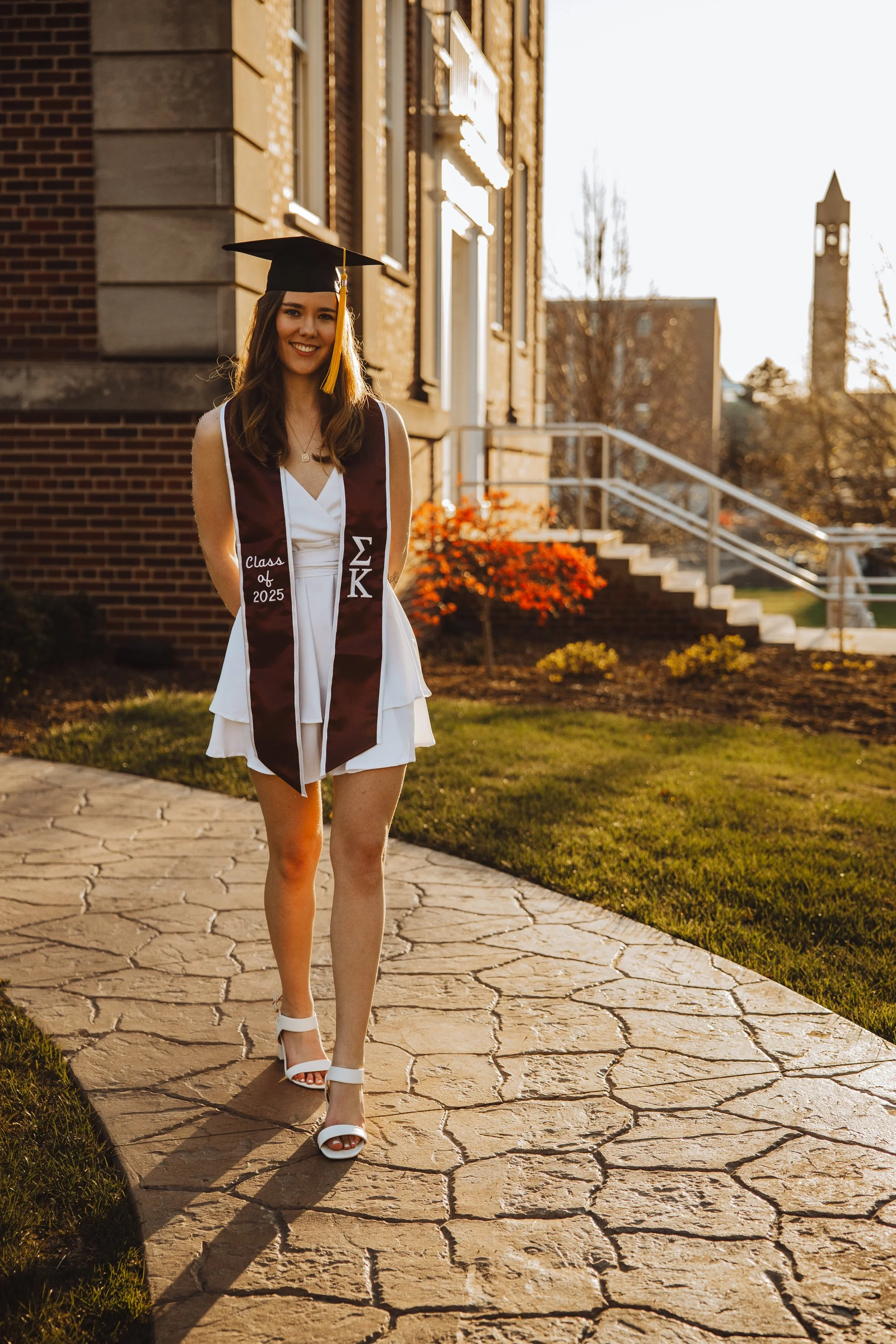 A young woman in a white dress and high heels standing outside on a stone pathway during sunset, wearing a maroon graduation stole with Greek letters and 'Class of 2025' embroidered on it, smiling at the camera.