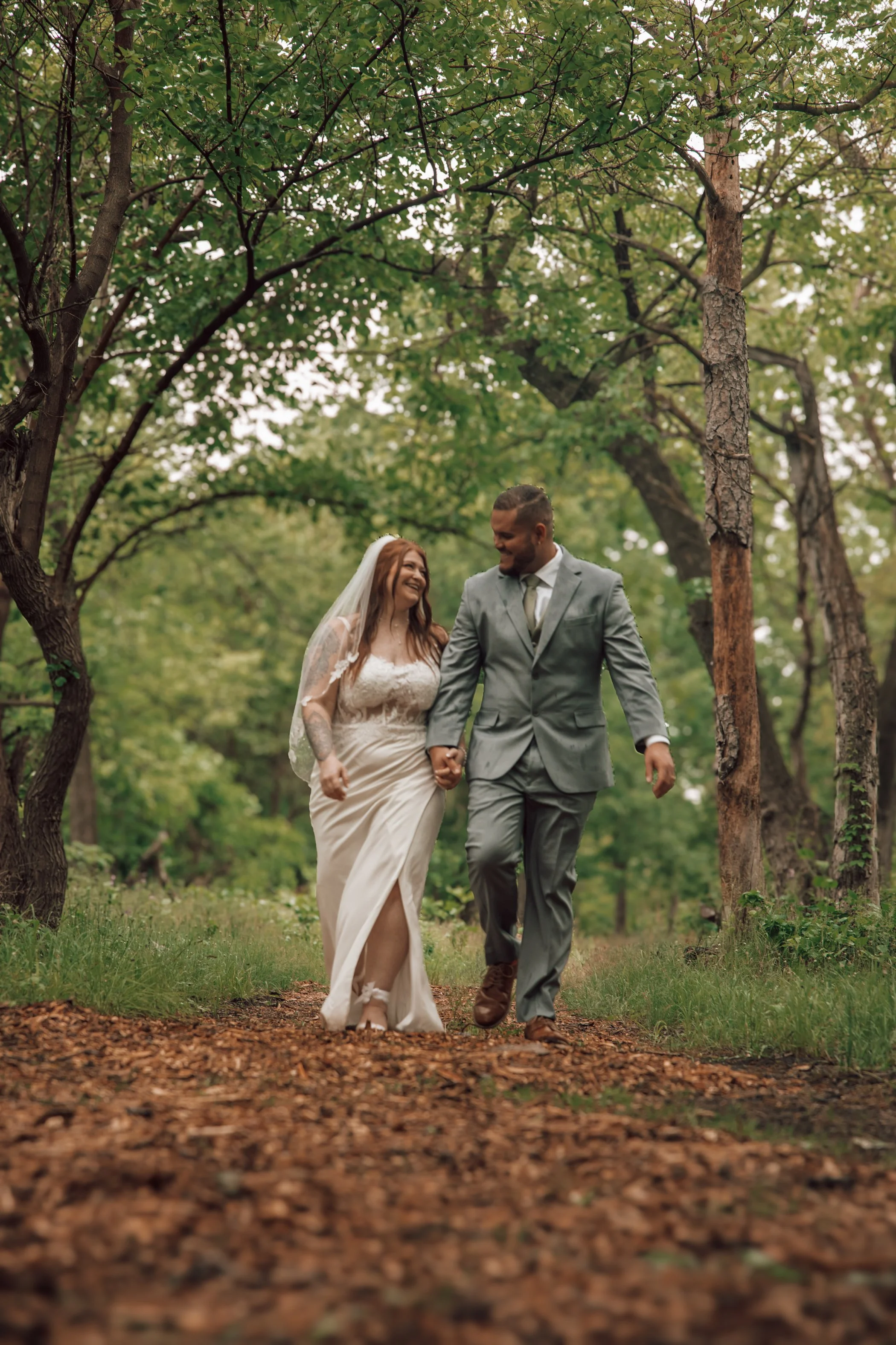 A happy couple in wedding attire walking hand in hand through a wooded area on a fall day.