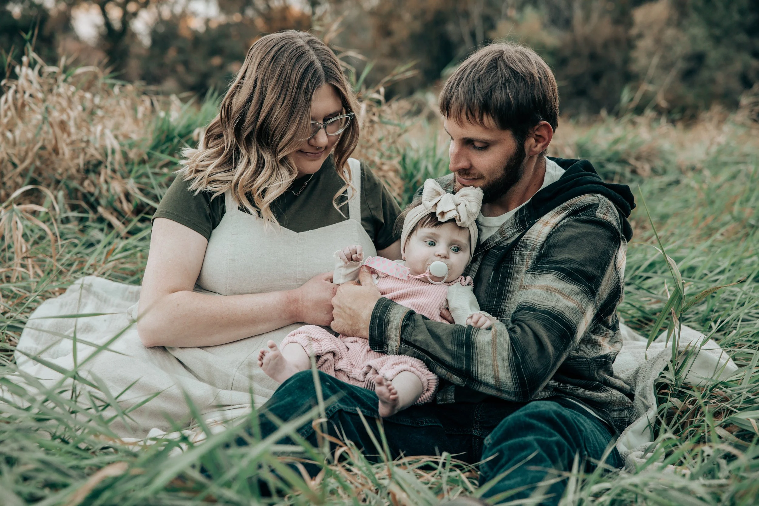 A family of three sitting outdoors on a blanket among tall grass, with a woman, a man, and a baby girl with a bow on her head, during fall.