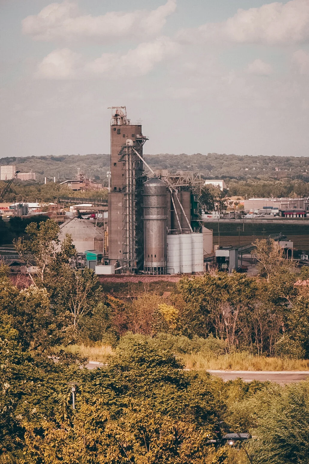 Industrial plant with tall buildings and silos, surrounded by trees and a distant landscape under a partly cloudy sky.