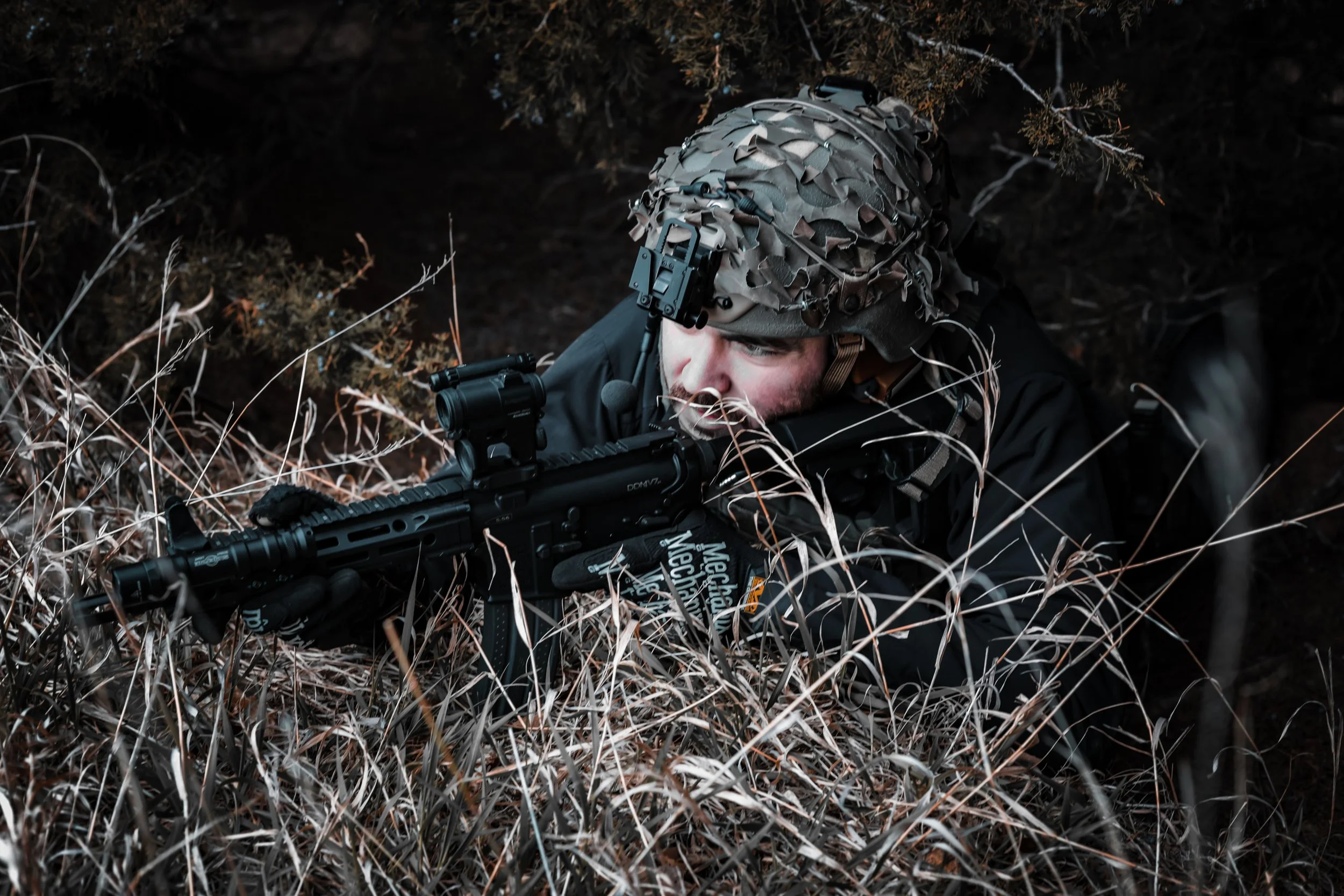 Soldier in camouflage gear lying on the ground among dry grass and bushes, aiming with a rifle.