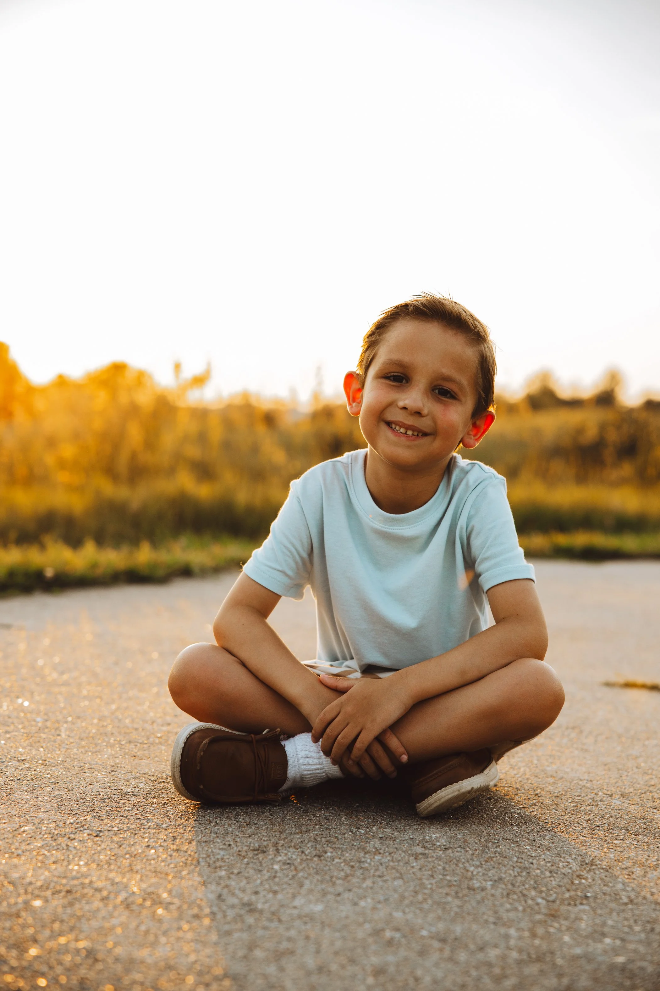 A young boy sitting cross-legged on the ground outdoors during golden hour with a smiling face, wearing a light blue t-shirt, brown shoes, and white socks, with a blurry sunset and autumn foliage in the background.
