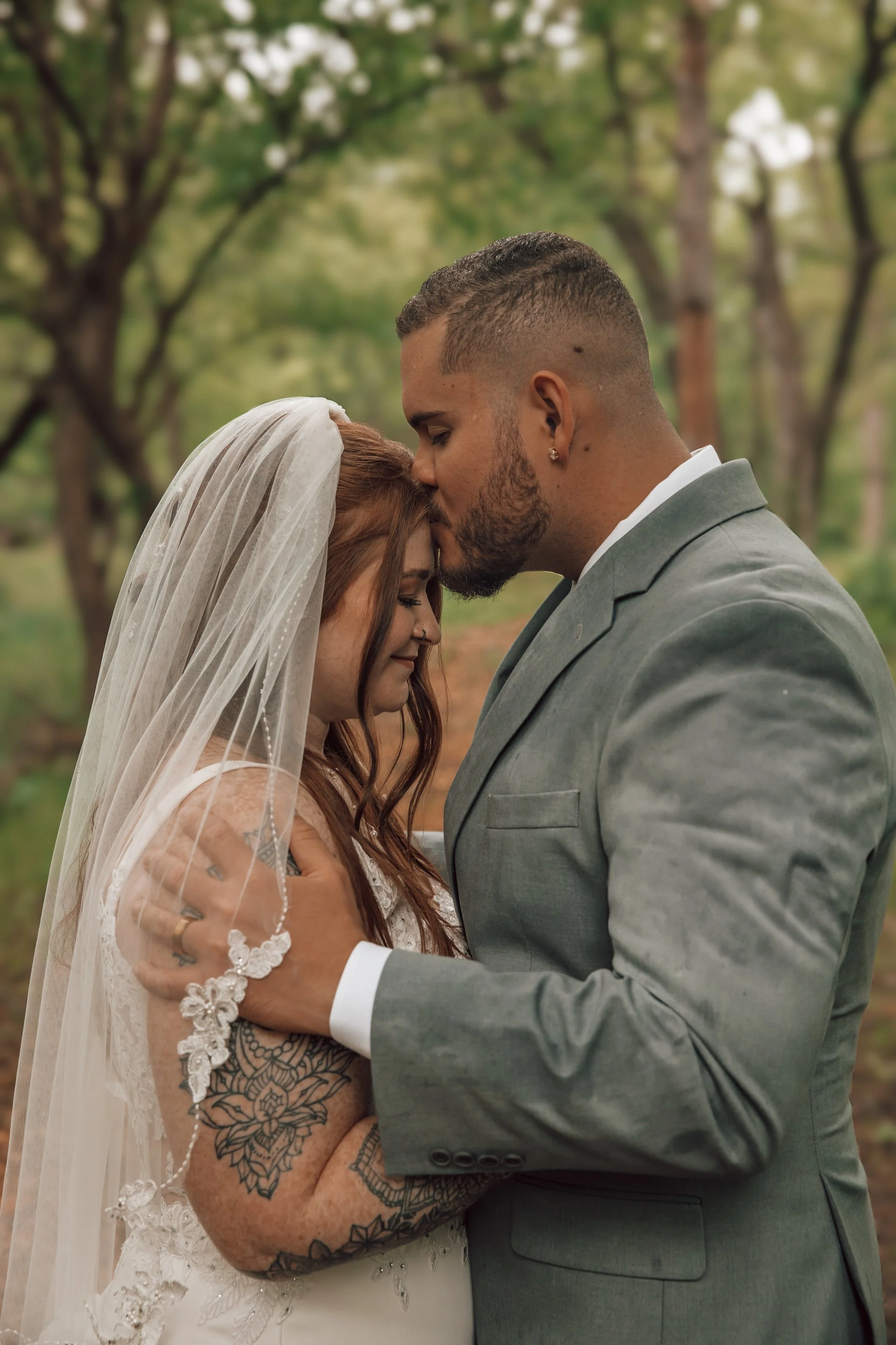 A couple is embracing in a forest, with the man kissing the woman's forehead. The woman is wearing a wedding veil and dress, and has tattoos on her arms. The man is dressed in a gray suit. They are holding hands and appear to be happy and intimate.