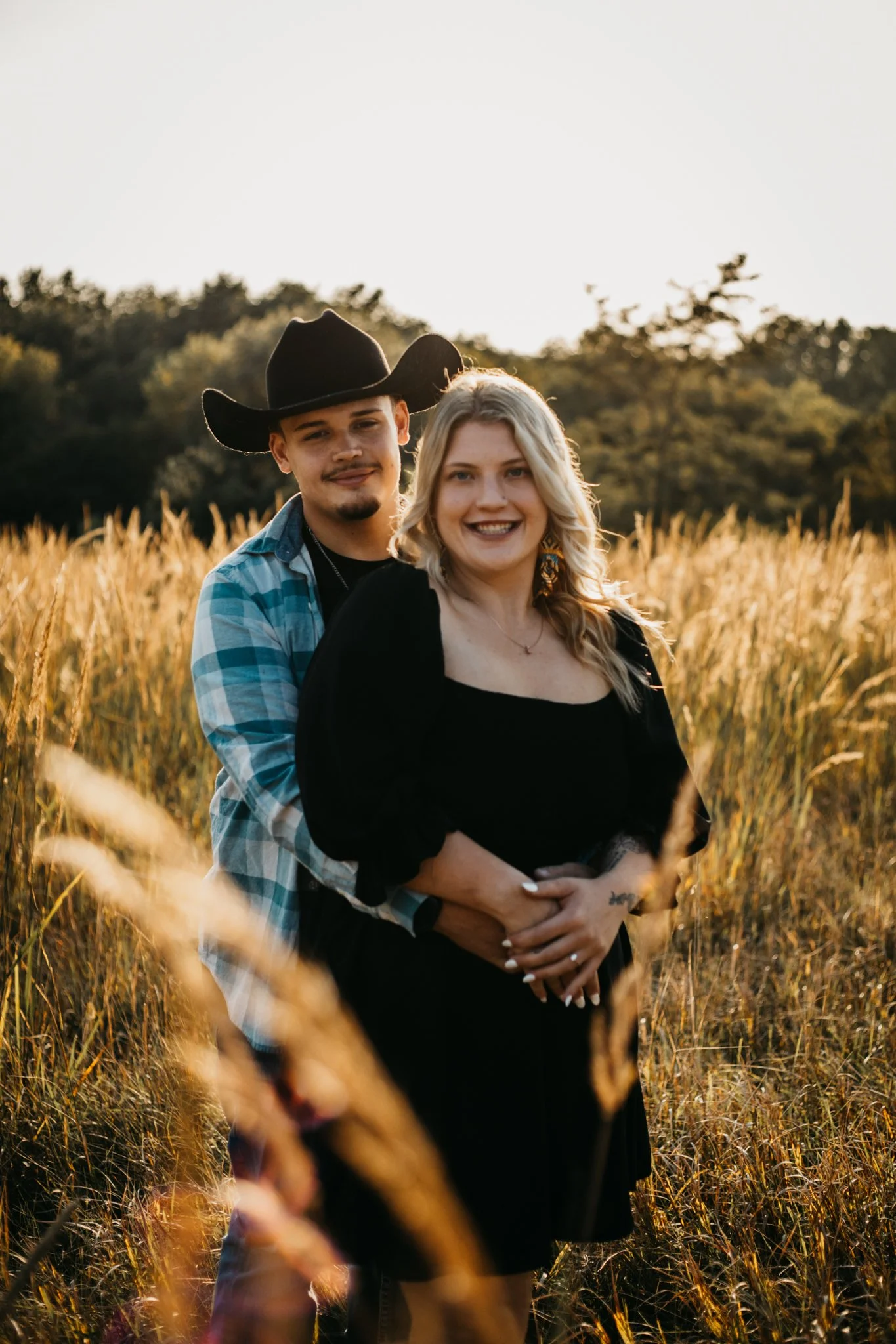 A couple standing in a field of tall grass during sunset, smiling and embracing each other, with trees in the background.