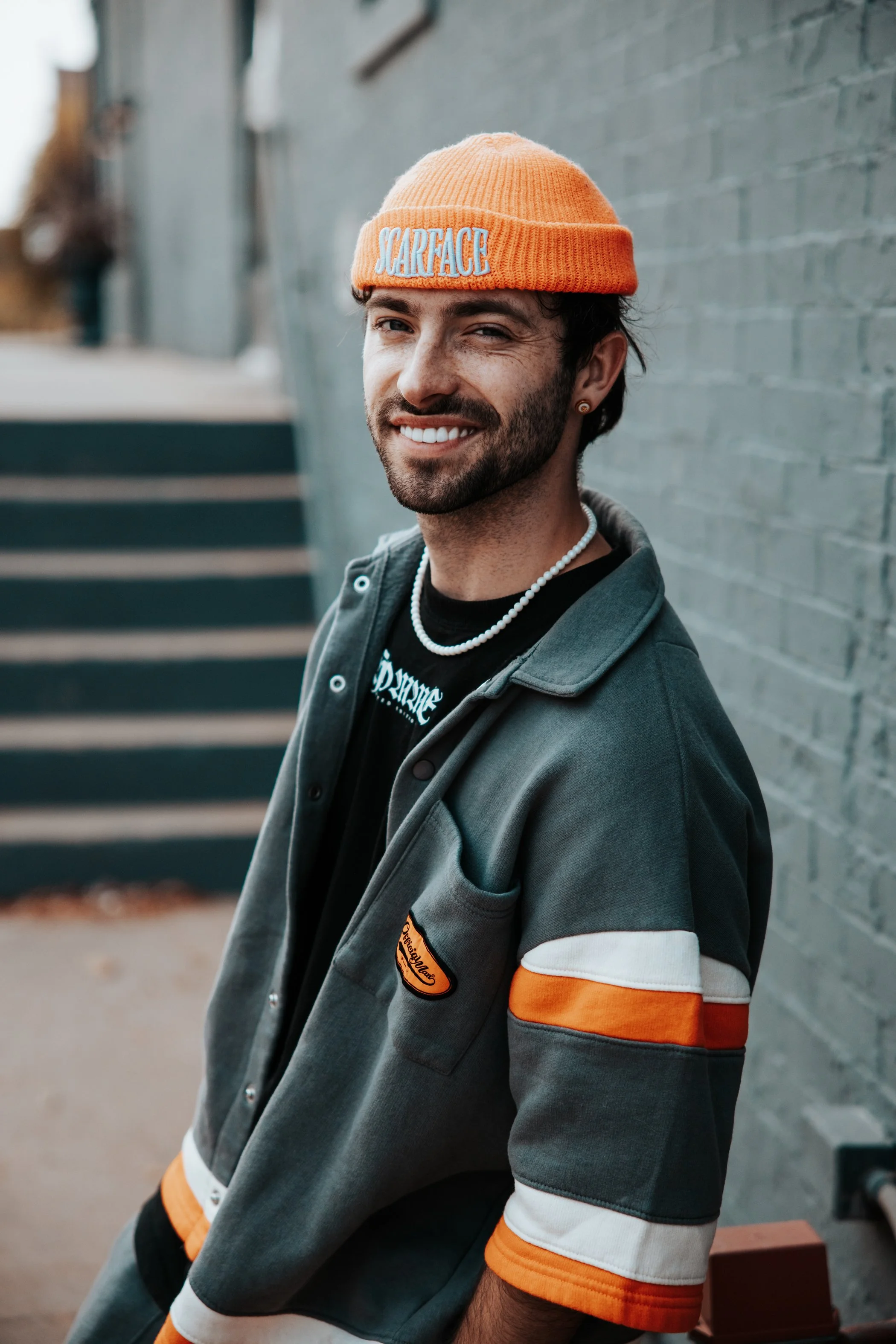 A smiling young man with dark hair and a beard wearing an orange beanie with the word "SCARFACE" embroidered on it, a pearl necklace, and a layered jacket with orange, black, and gray colors, standing outdoors against a gray brick wall background.