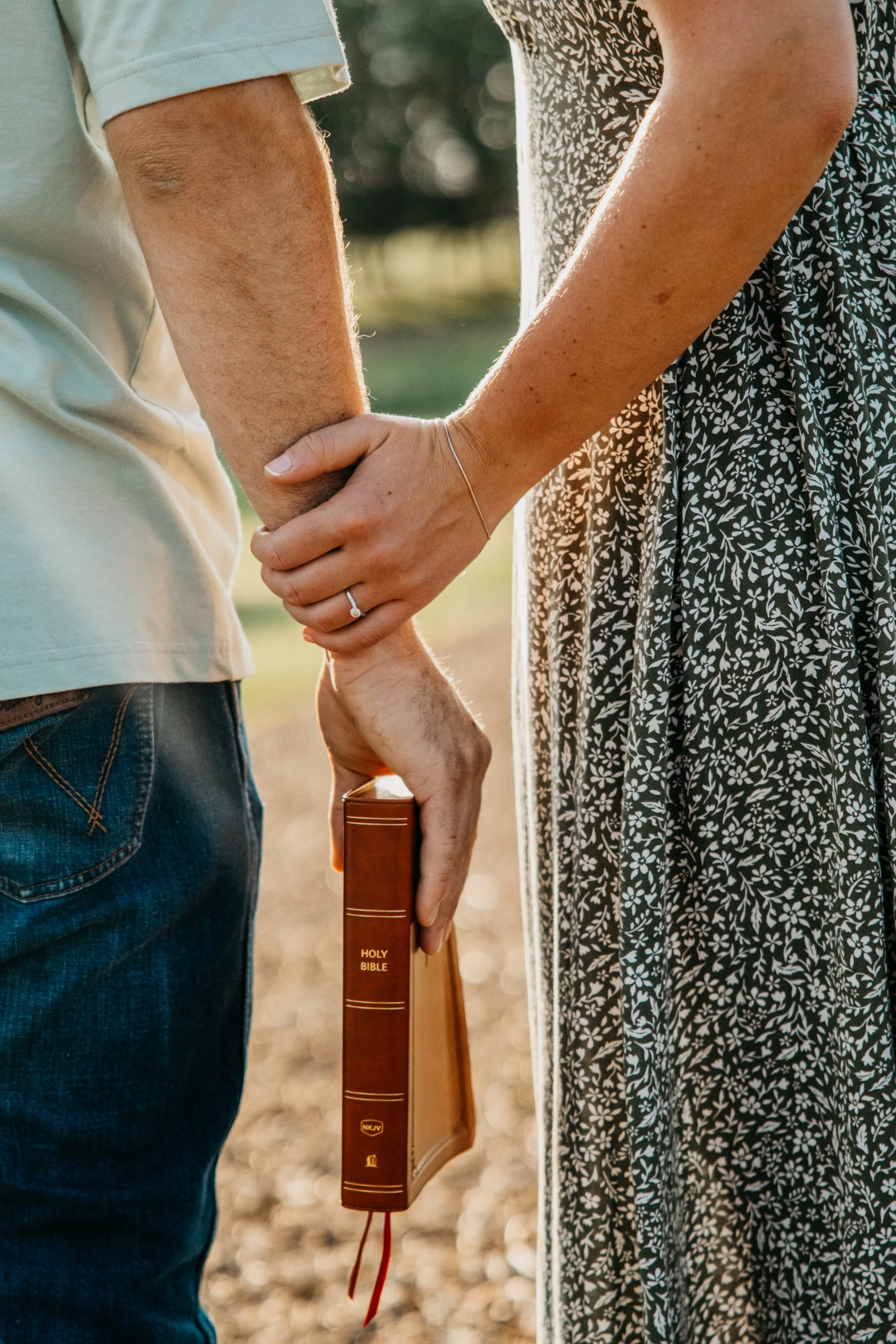 Close-up of a couple holding hands, with the woman holding a Bible, in a natural outdoor setting during golden hour.