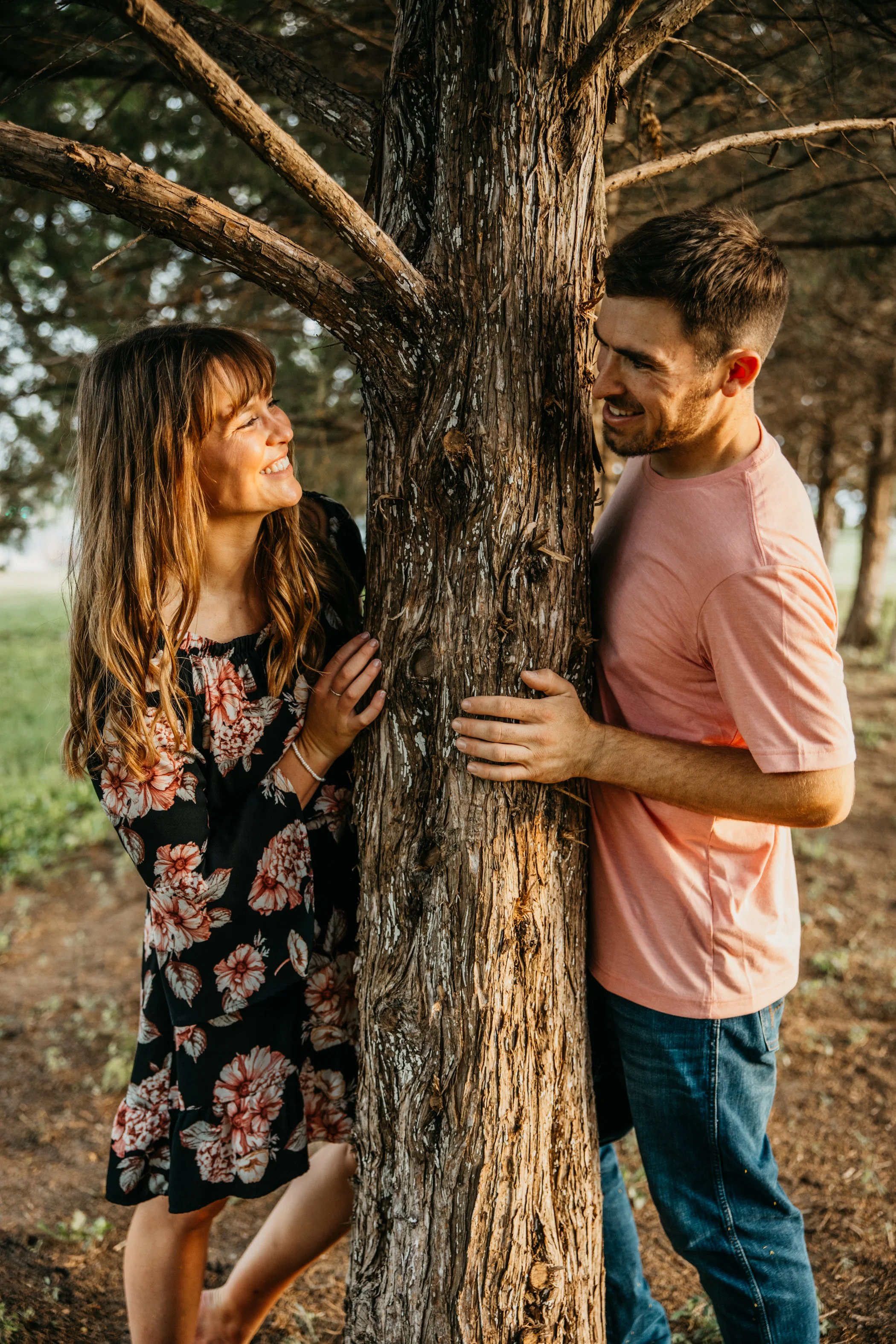A man and woman are smiling and gazing at each other while standing on either side of a tree. They are outdoors in a wooded area, with the woman's hand on the tree and the man's hand holding the tree trunk. The woman has long, wavy reddish hair and i